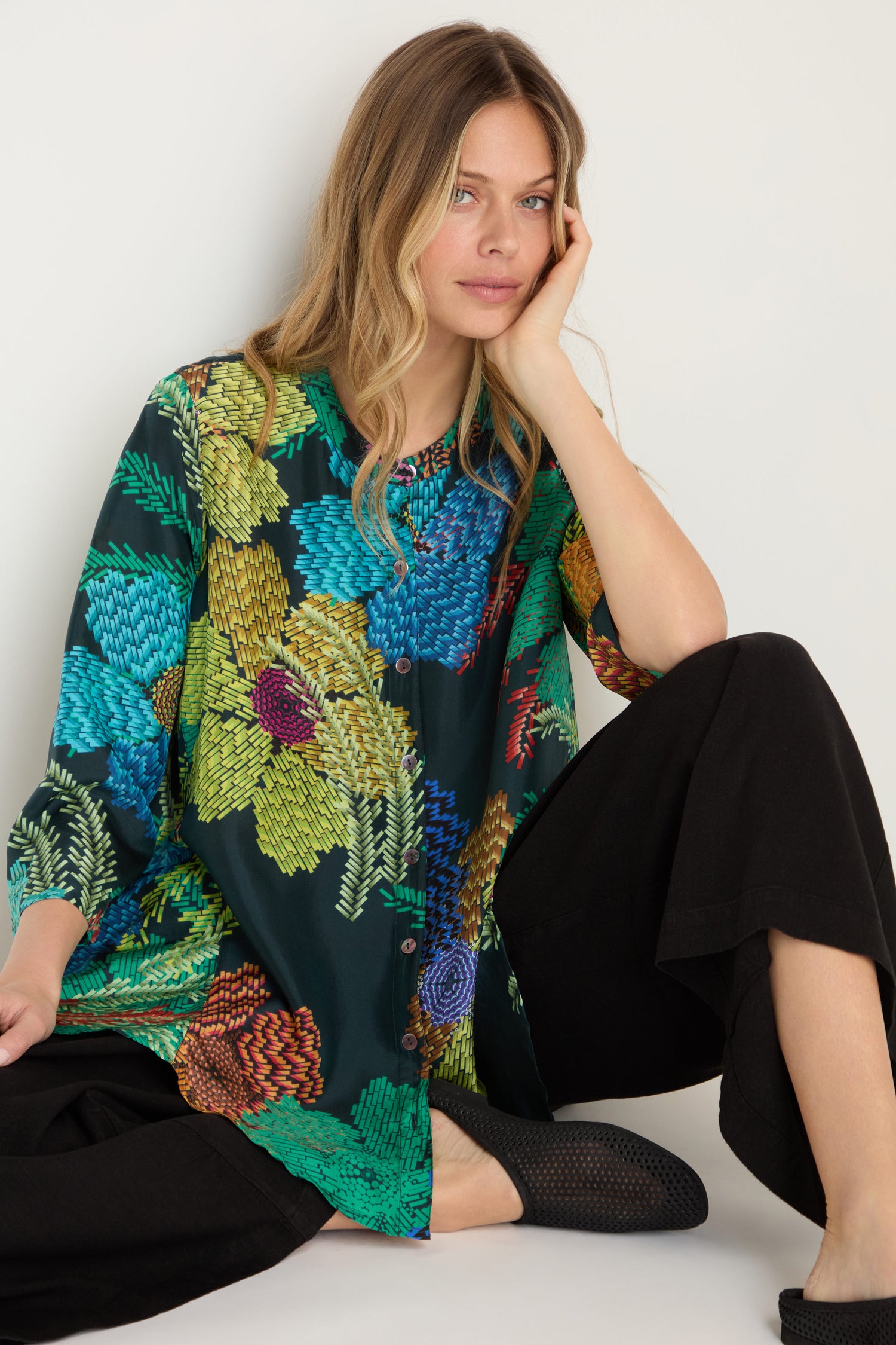 A woman with long blonde hair wears the Pixelated Petals Print Silk Shirt and black pants as she sits on the floor, resting her head on her hand against a plain background.