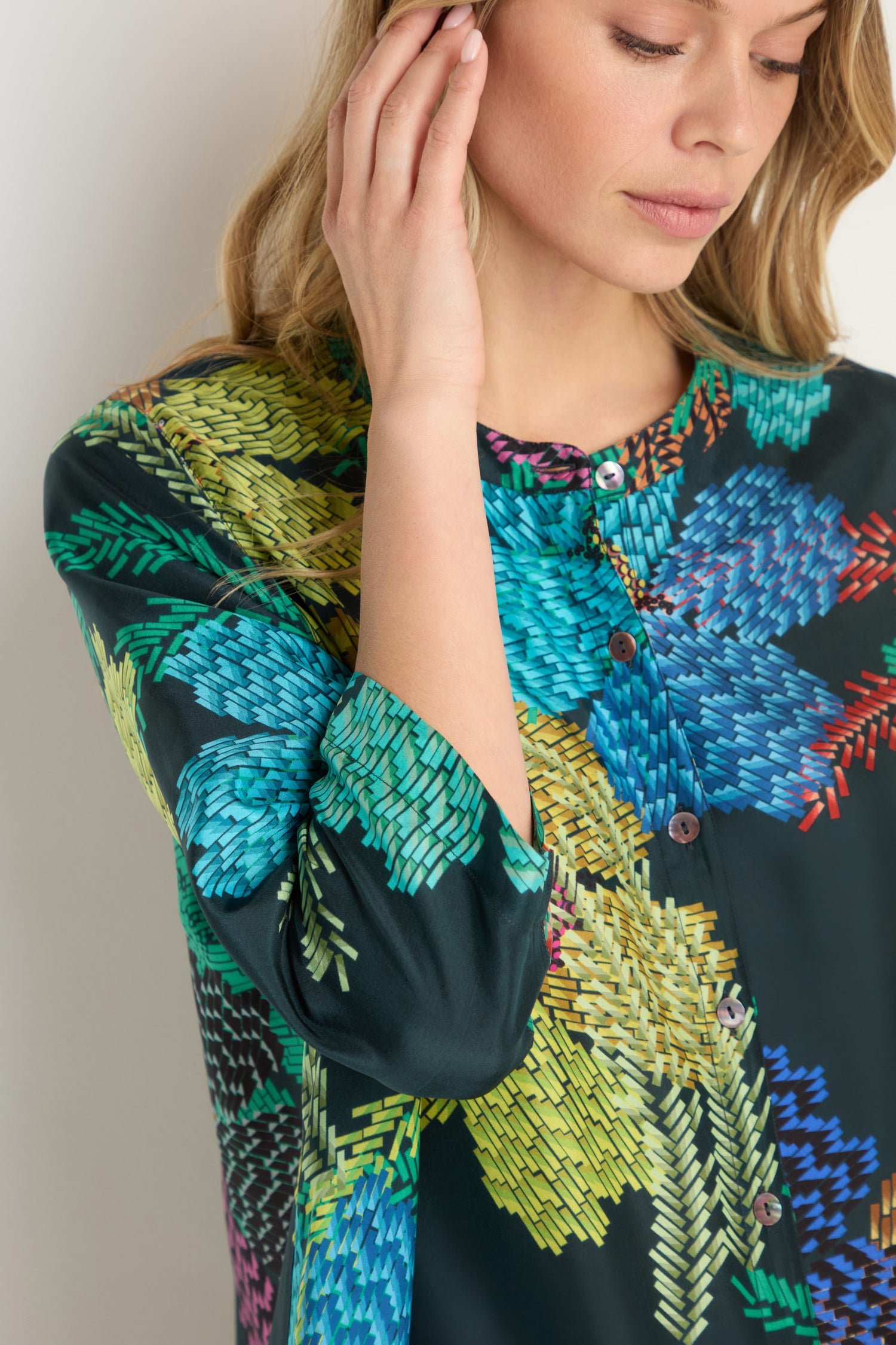 A woman wearing the Pixelated Petals Print Silk Shirt, a dark blouse adorned with woven pixel floral designs, touches her face with one hand.