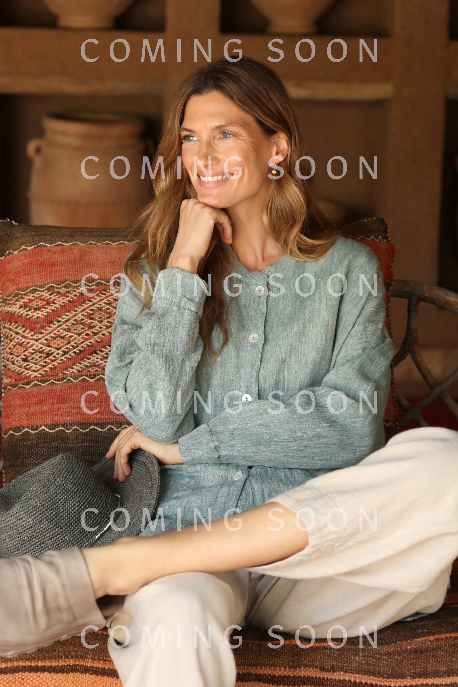 A woman with long hair sits on a patterned sofa, wearing the Crinkle Linen Cross Dye Shirt in light blue and light pants, smiling with her hand on her chin. The words "COMING SOON GRT6777-TXCD" appear on the image.