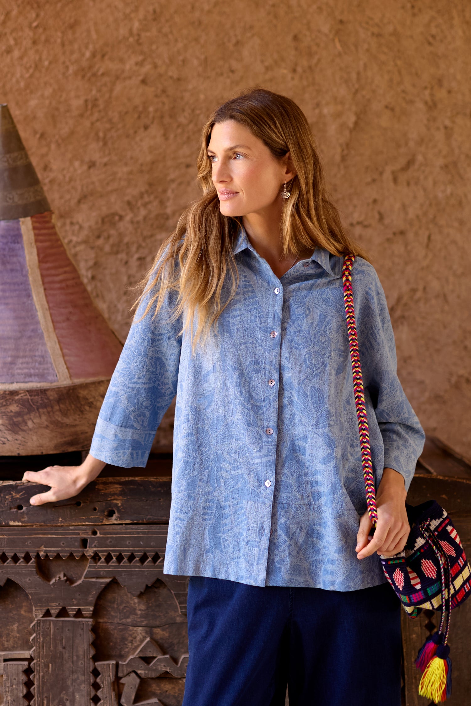 A woman stands indoors, wearing a Floral Jacquard Cotton Shirt and dark pants, with her hand on a carved wooden surface and a colorful GRT6856-FJC bag over her shoulder.