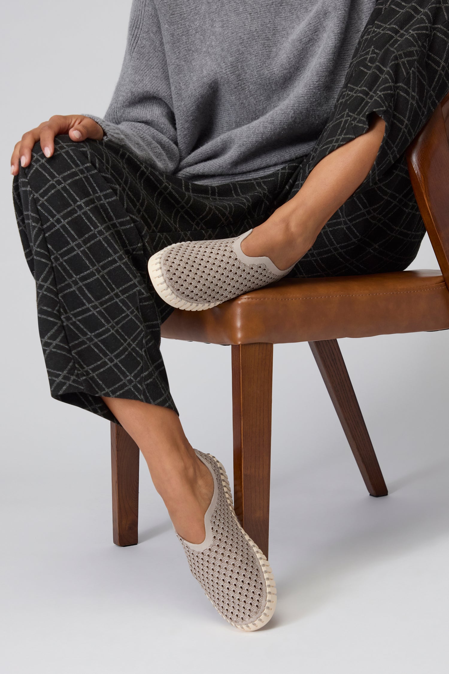 A person seated on a brown chair wearing gray plaid pants, a gray sweater, and Grey Tulip Slip On Shoes. Only the lower body is visible, highlighting sustainable shoes inspired by Scandinavian design.