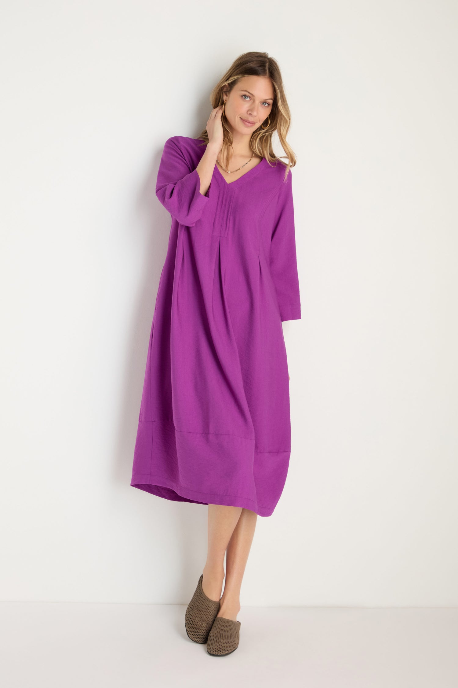 A woman stands against a white wall wearing the Textured Viscose Bubble Dress in orchid purple, a loose-fitting, long-sleeved midi dress, paired with brown slip-on shoes.