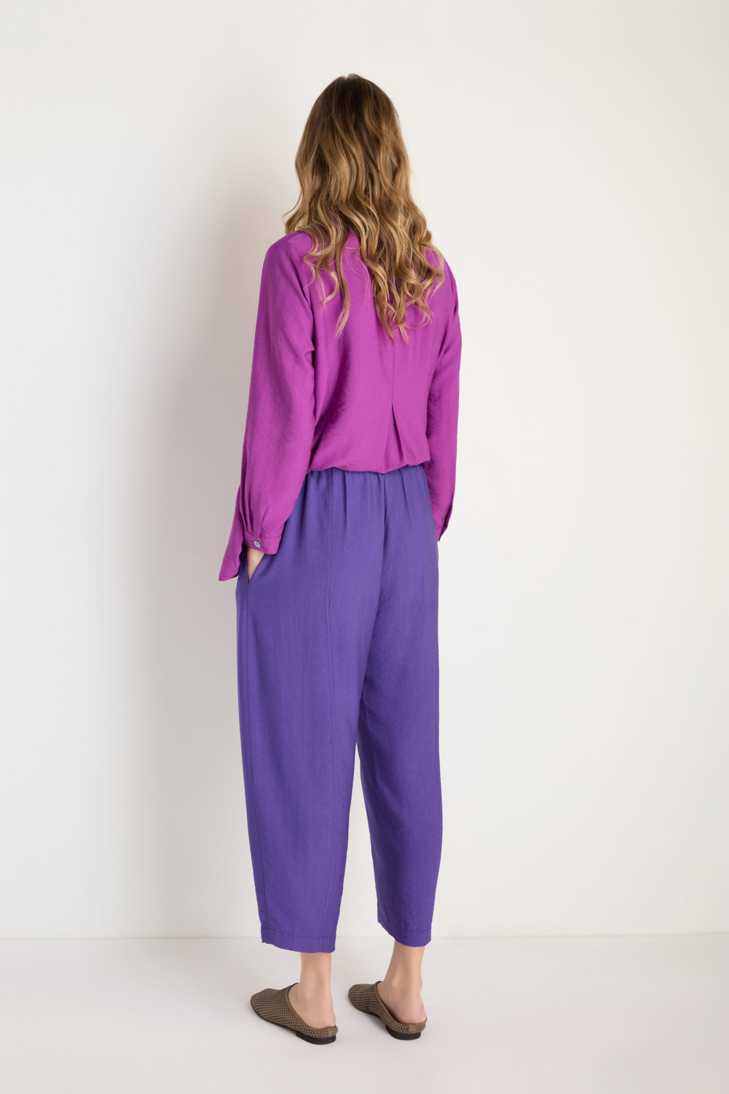 A woman with wavy hair, facing away, wears a purple long-sleeve top and cropped Textured Viscose Bubble Trouser paired with flat shoes against a plain white background.