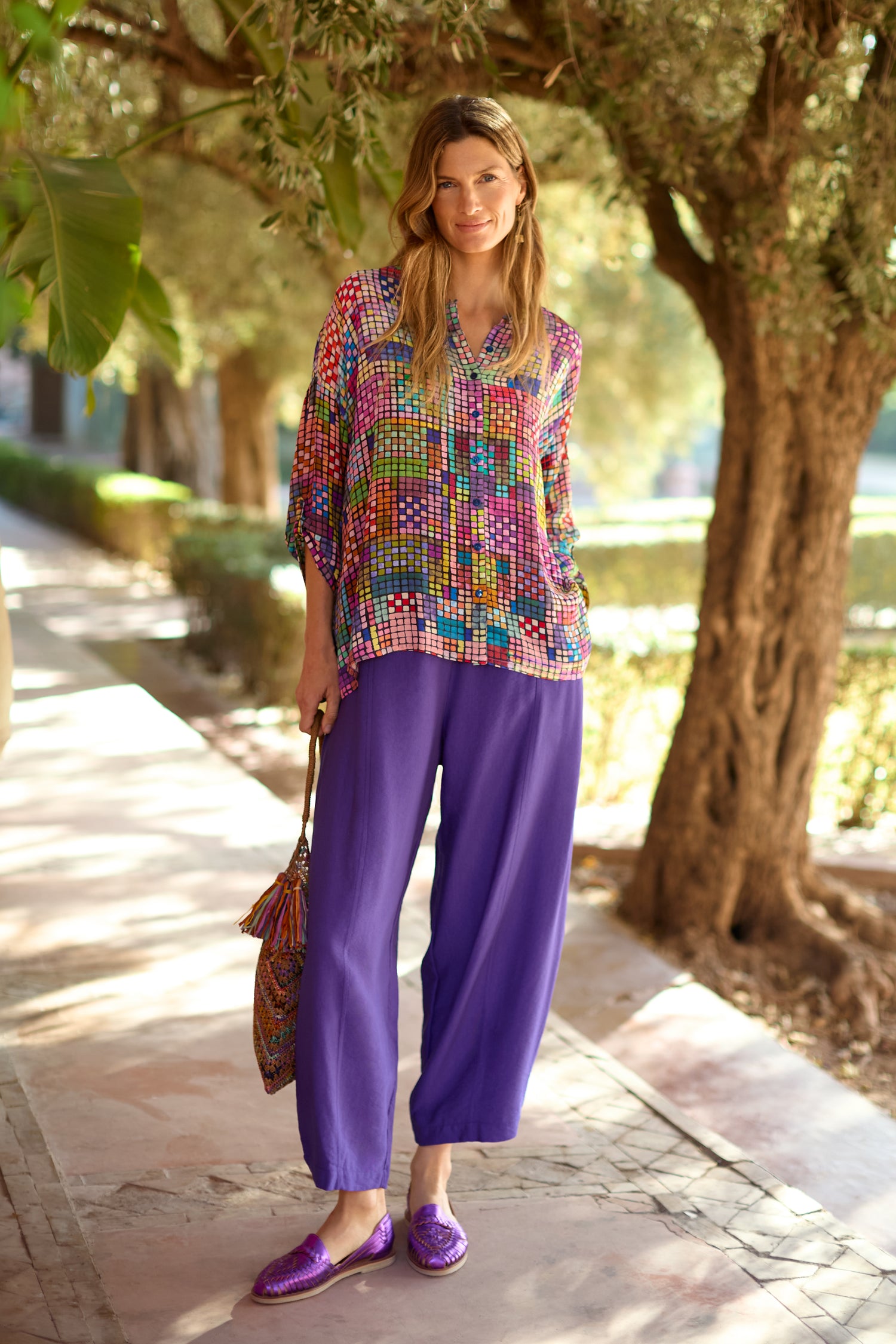 A woman stands outdoors on a stone path, wearing the Textured Viscose Bubble Trouser in purple, a colorful patterned top, matching slip-on shoes, and holding a coordinating bag. Trees and greenery fill the background.