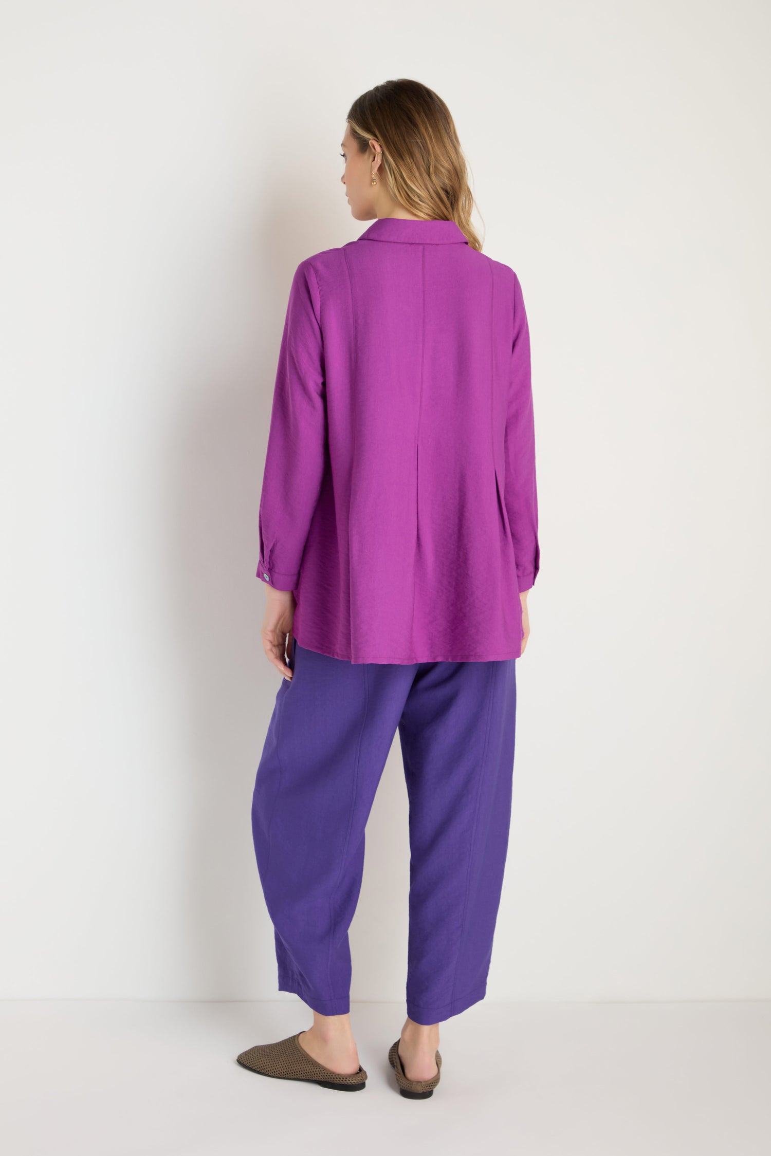 A person stands facing away against a plain white background, wearing a long-sleeved magenta Textured Viscose Pleat Shirt, loose purple pants, and brown slip-on shoes.