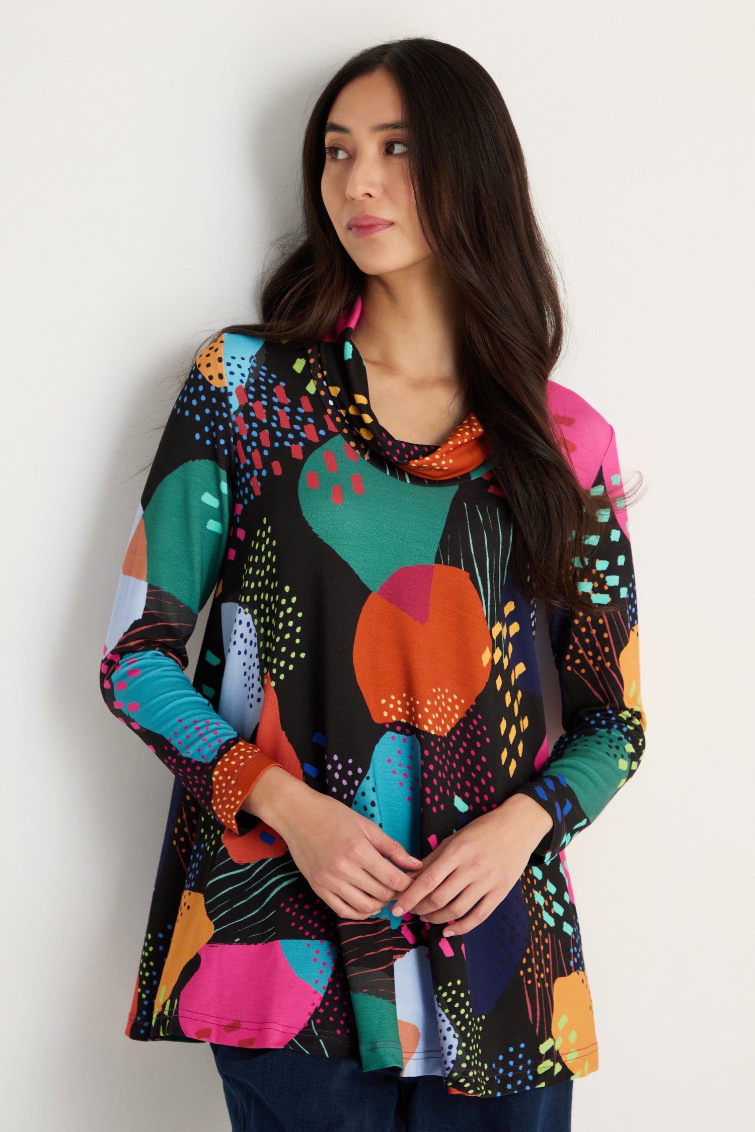 A woman stands against a white wall wearing the Mark Making Print Jersey Tunic with long sleeves, a cowl neck, and dark pants.