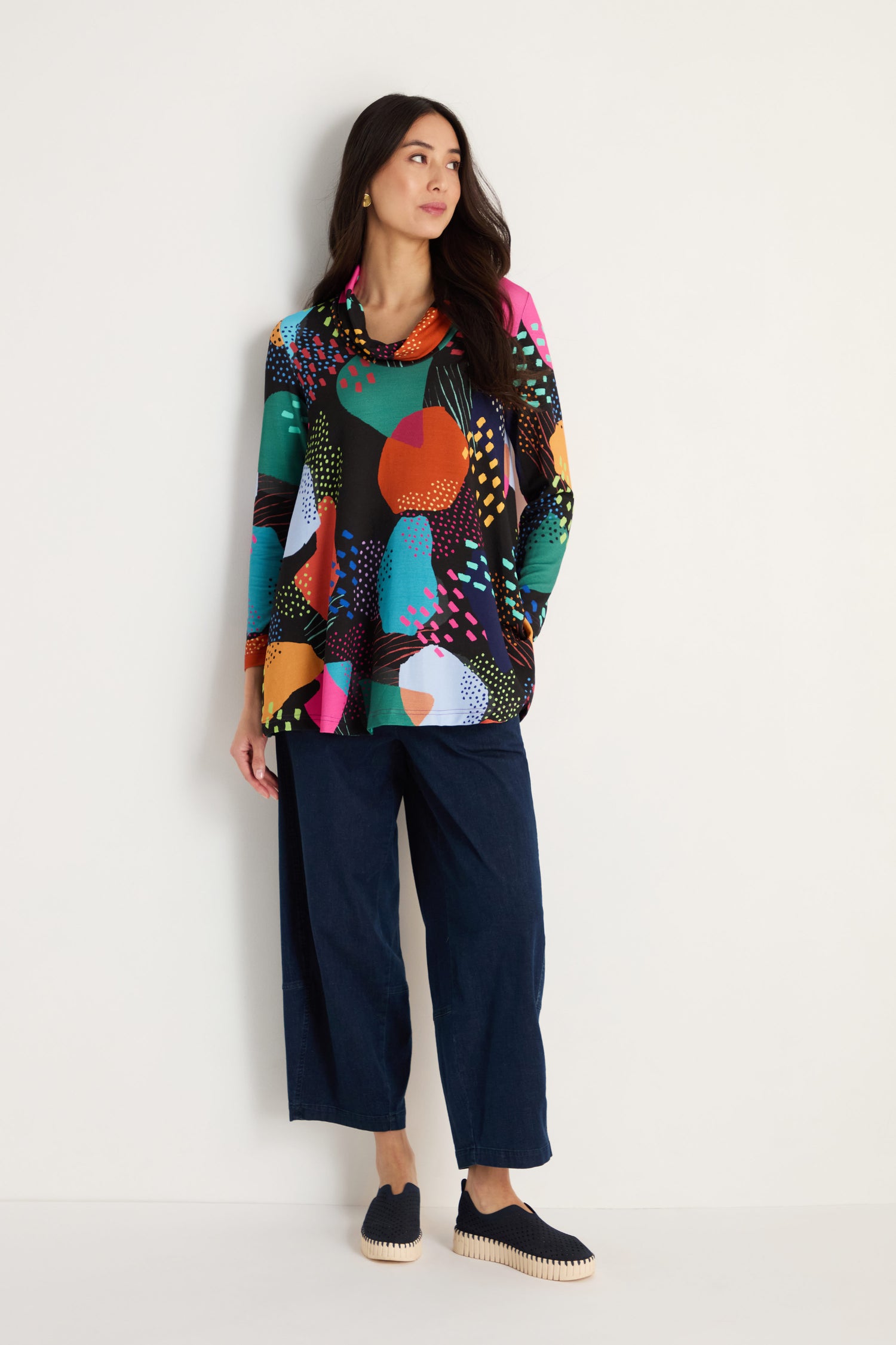 A woman stands against a white wall wearing the Mark Making Print Jersey Tunic, wide-leg dark pants, and black slip-on shoes.