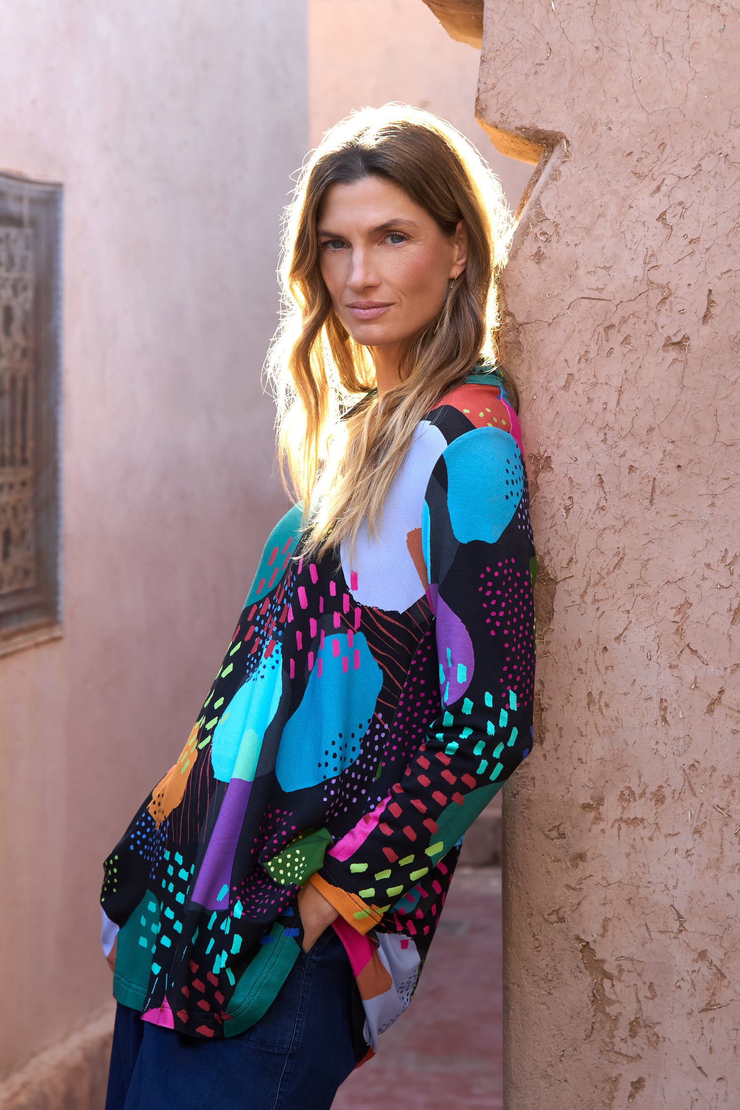 A woman with long hair wears the Colour Play Print Jersey Tunic and blue pants as she leans against a textured wall outdoors.