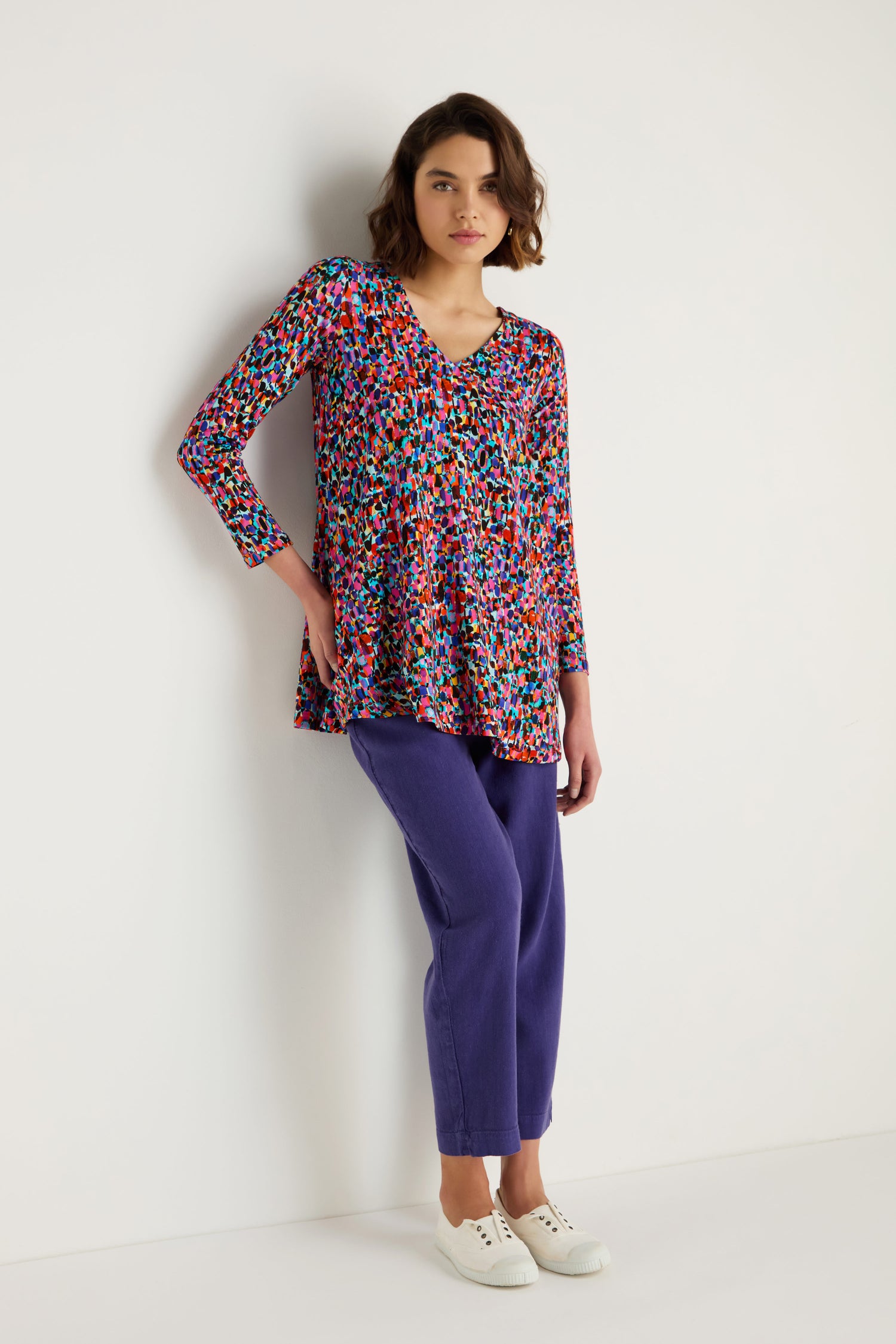 A woman stands against a white wall wearing the Painted Spots Jersey Tunic, paired with purple pants and white sneakers.