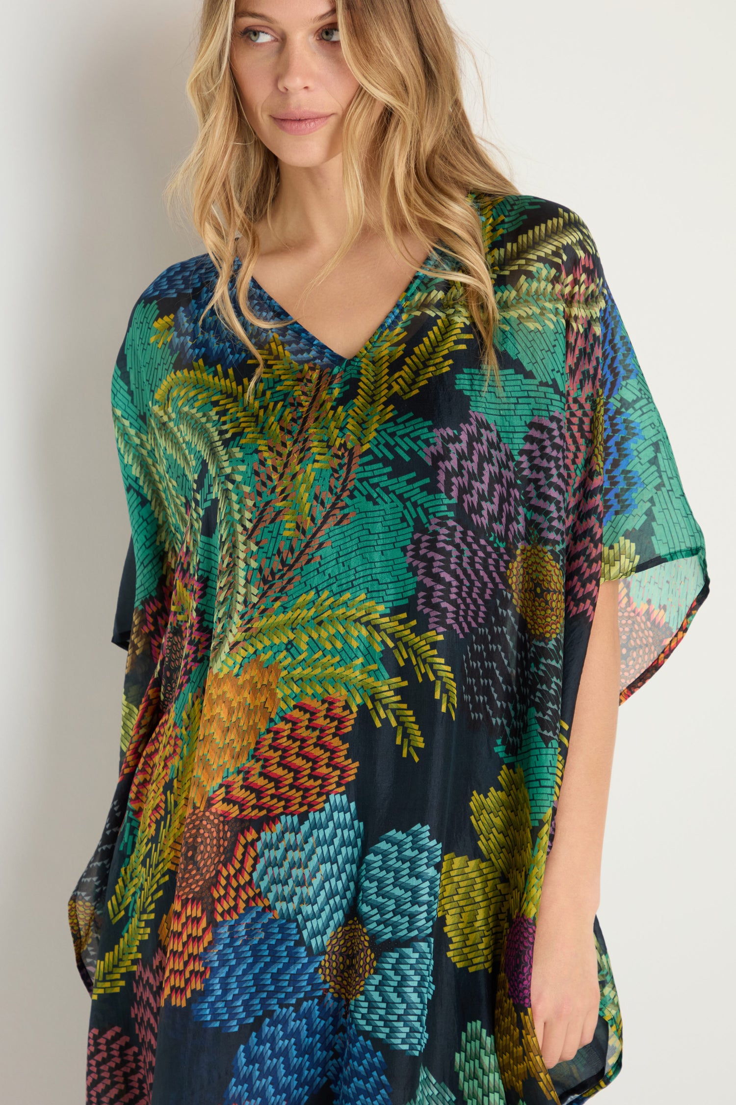 A woman wears the Pixelated Petals Silk V-Neck Kaftan, featuring a colorful pixelated floral print.