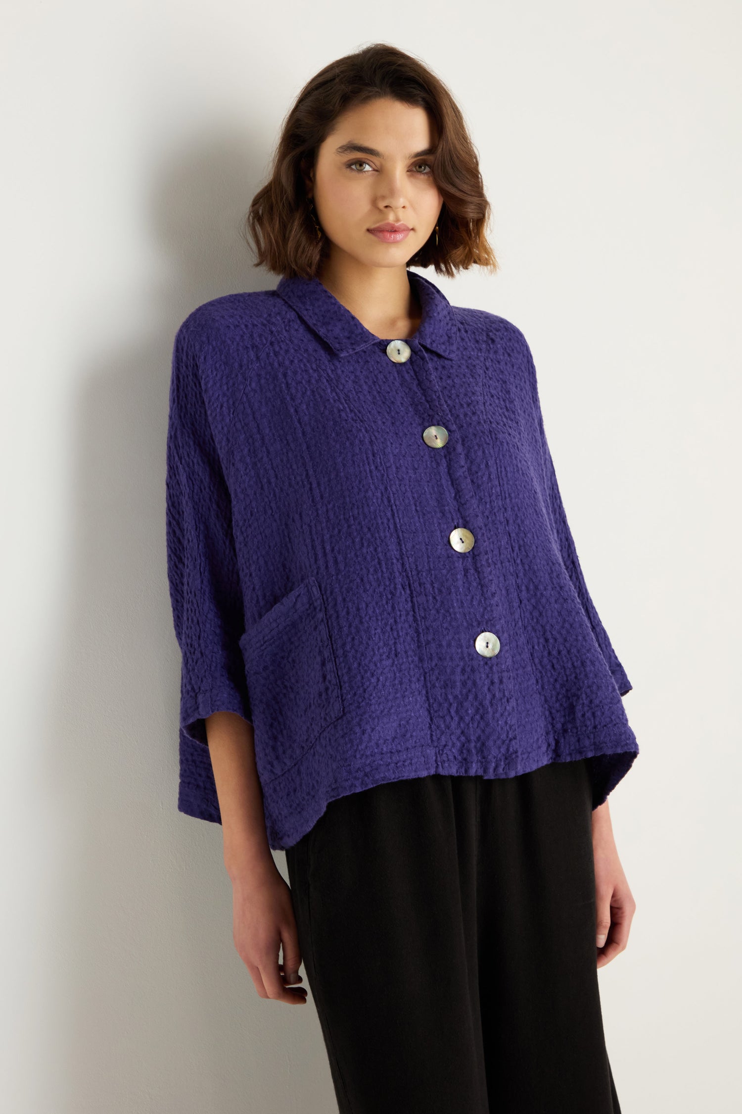 A woman stands against a white wall wearing the Waffle Linen Boxy Jacket in textured purple with large buttons and black pants.