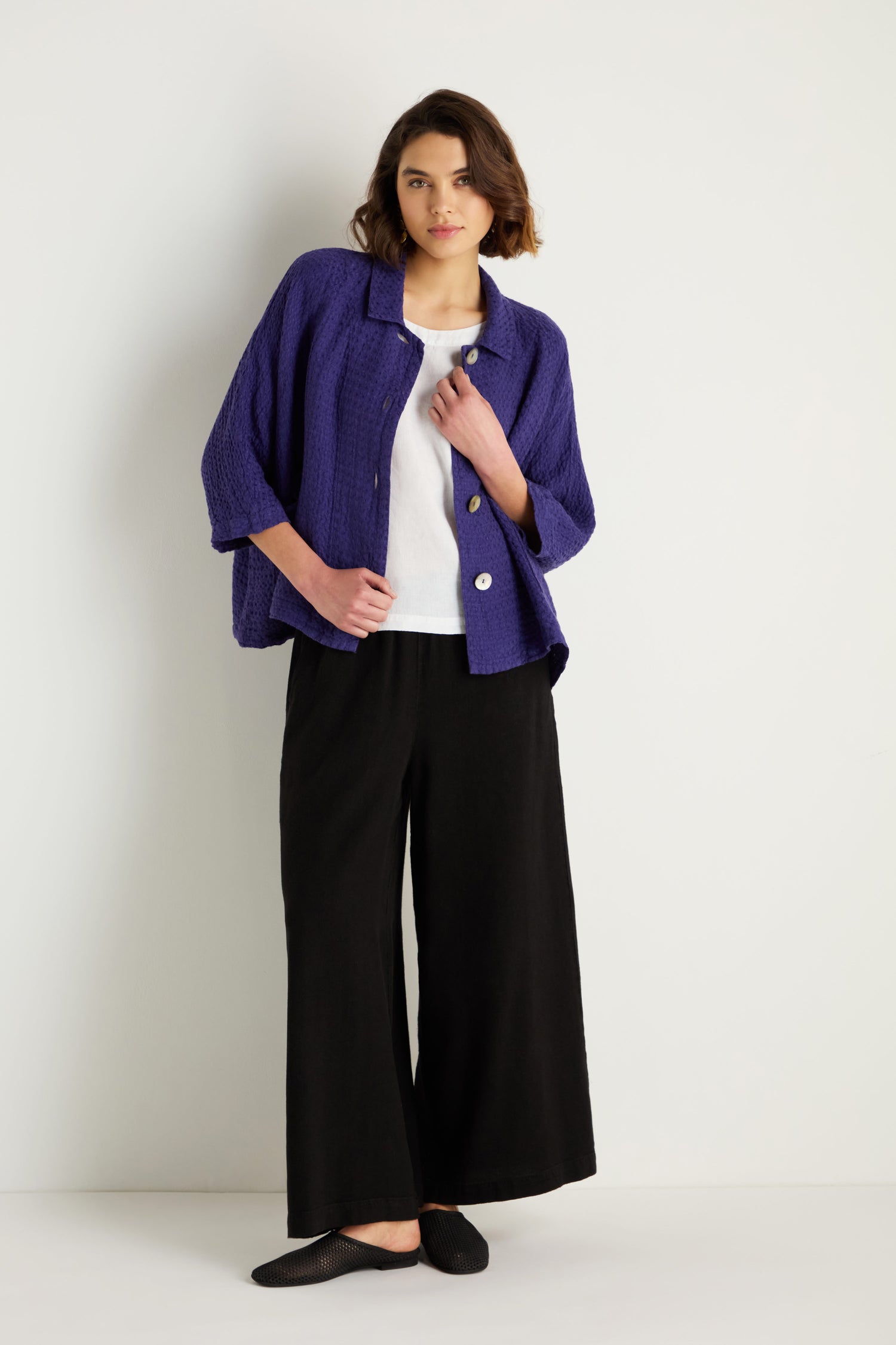 A person stands against a white wall, wearing the Waffle Linen Boxy Jacket in purple over a white shirt, with black wide-leg pants and black shoes.