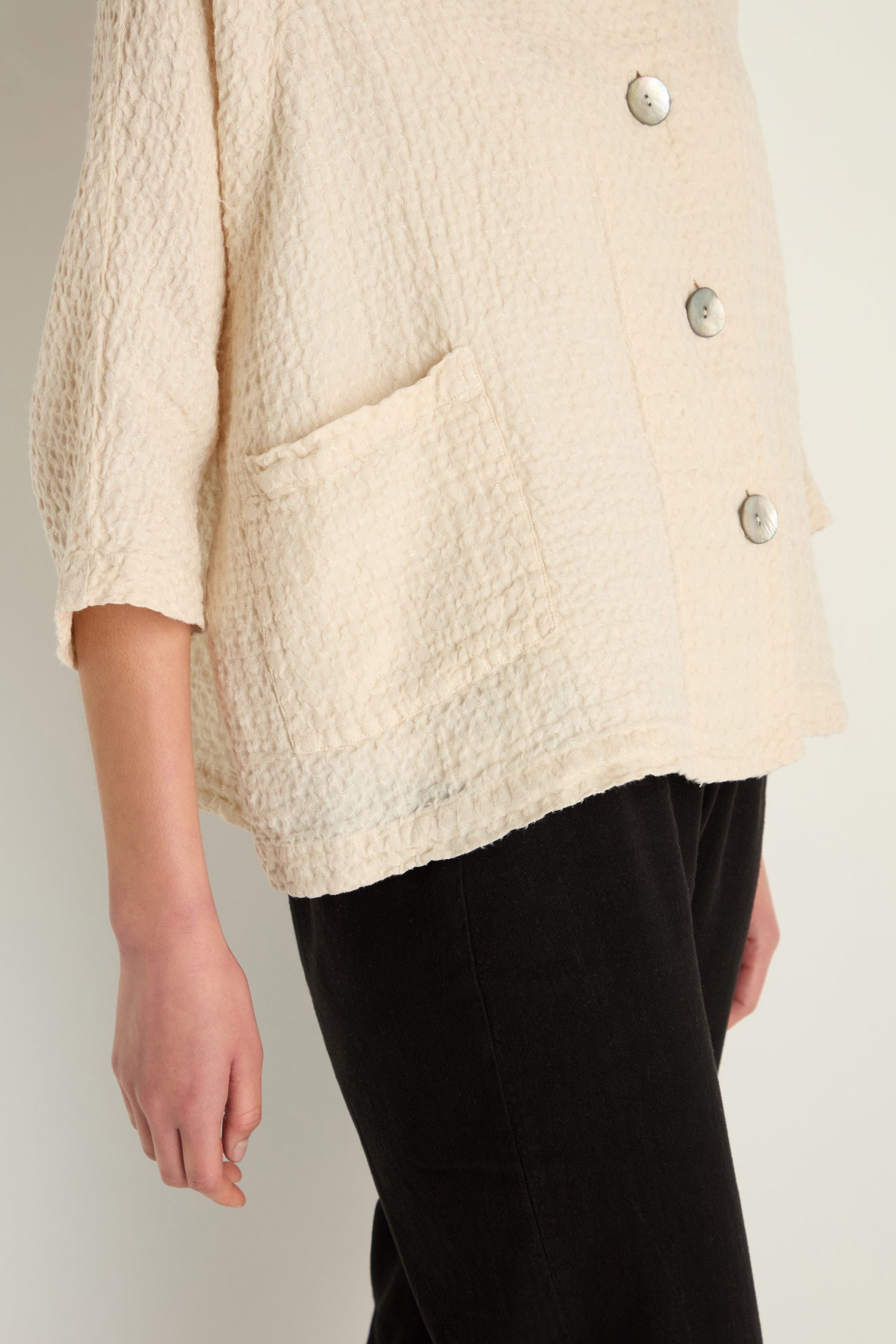 A person wearing the Waffle Linen Boxy Jacket—a textured, cream-colored, oversized linen jacket with large buttons and a front pocket—paired with black pants.