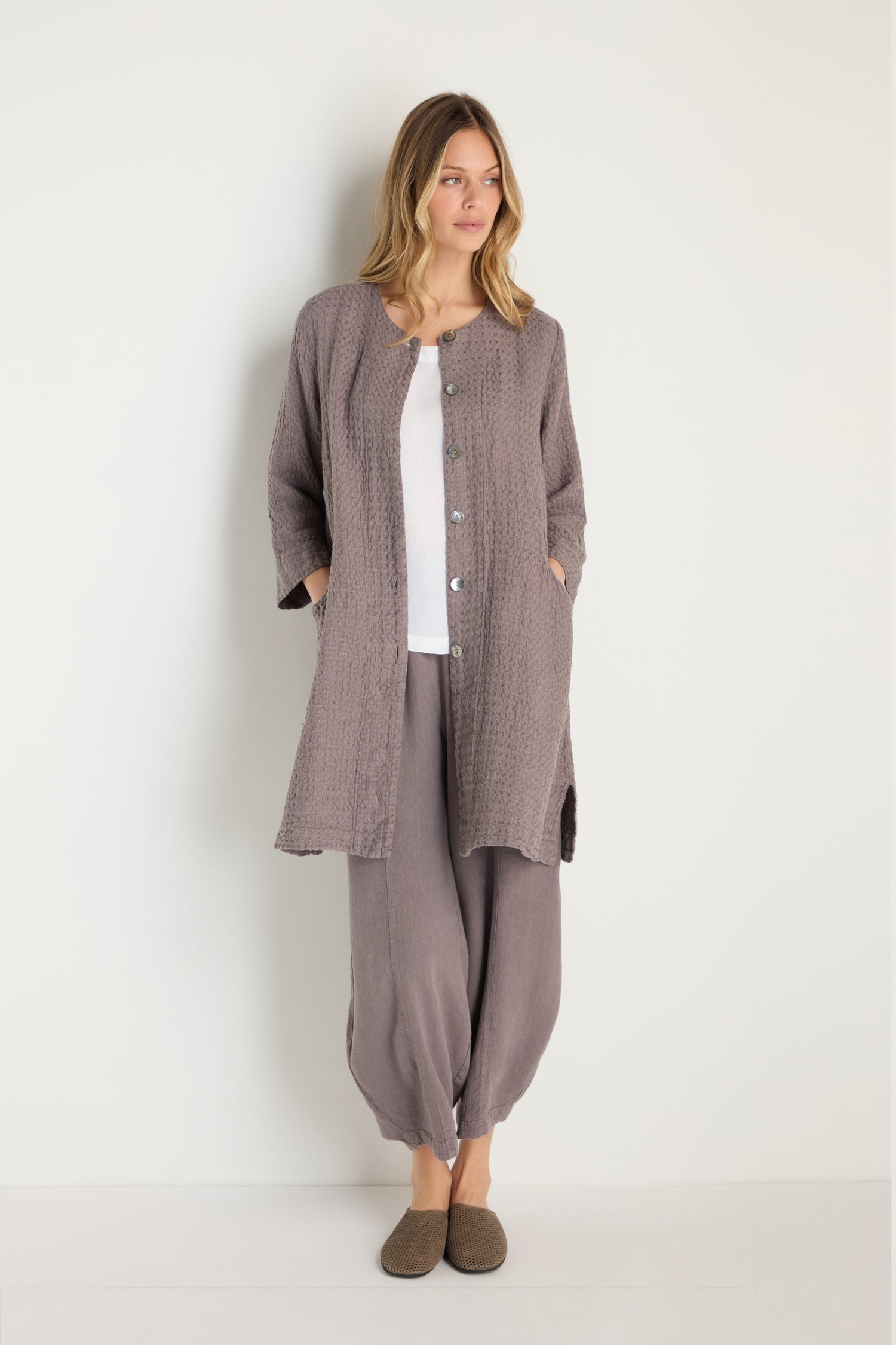 A woman stands against a white wall wearing the Waffle Linen Long Jacket in mauve with matching loose pants, a white top, and brown slippers. Hands in pockets, she gazes to the side—an effortlessly chic wardrobe essential.