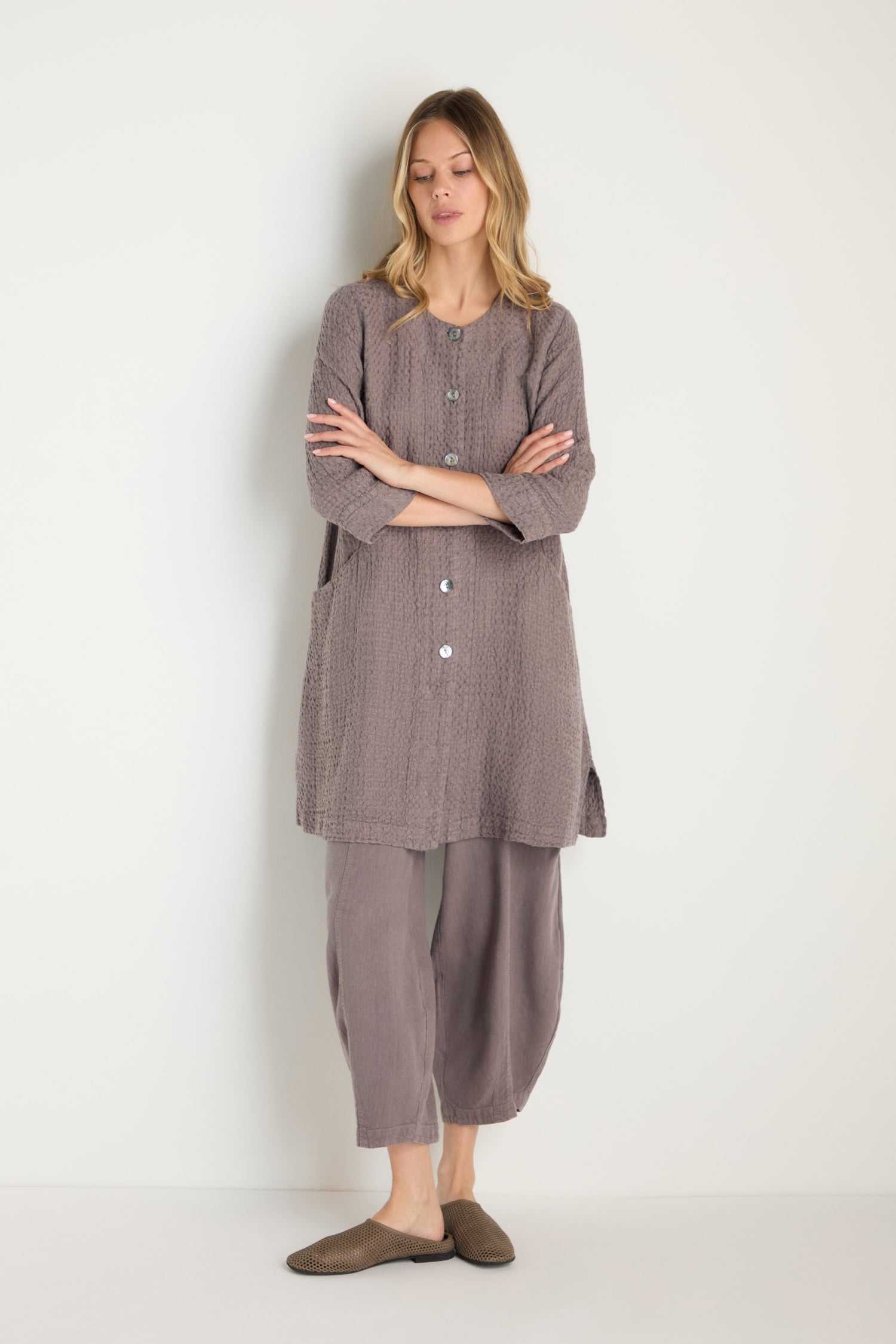 A woman stands against a plain background wearing the Waffle Linen Long Jacket in muted gray tones paired with matching pants and brown slip-on shoes—effortless, breathable wardrobe essentials.