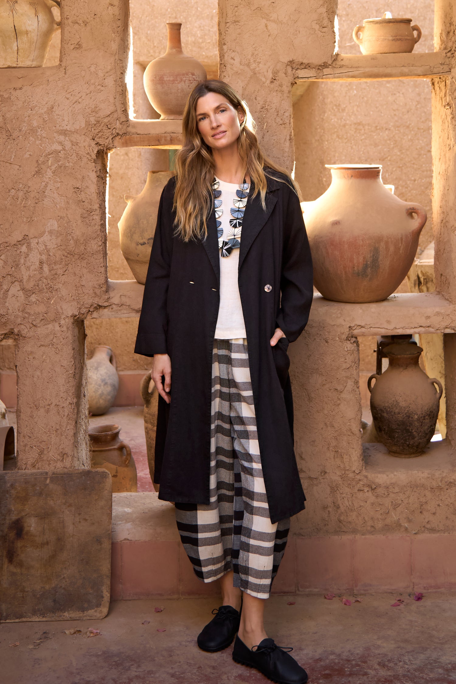 A woman stands indoors in a rustic setting, wearing the Twisted Linen Relaxed Coat, a white printed shirt, and black-and-white plaid pants, surrounded by large clay pots on shelves.