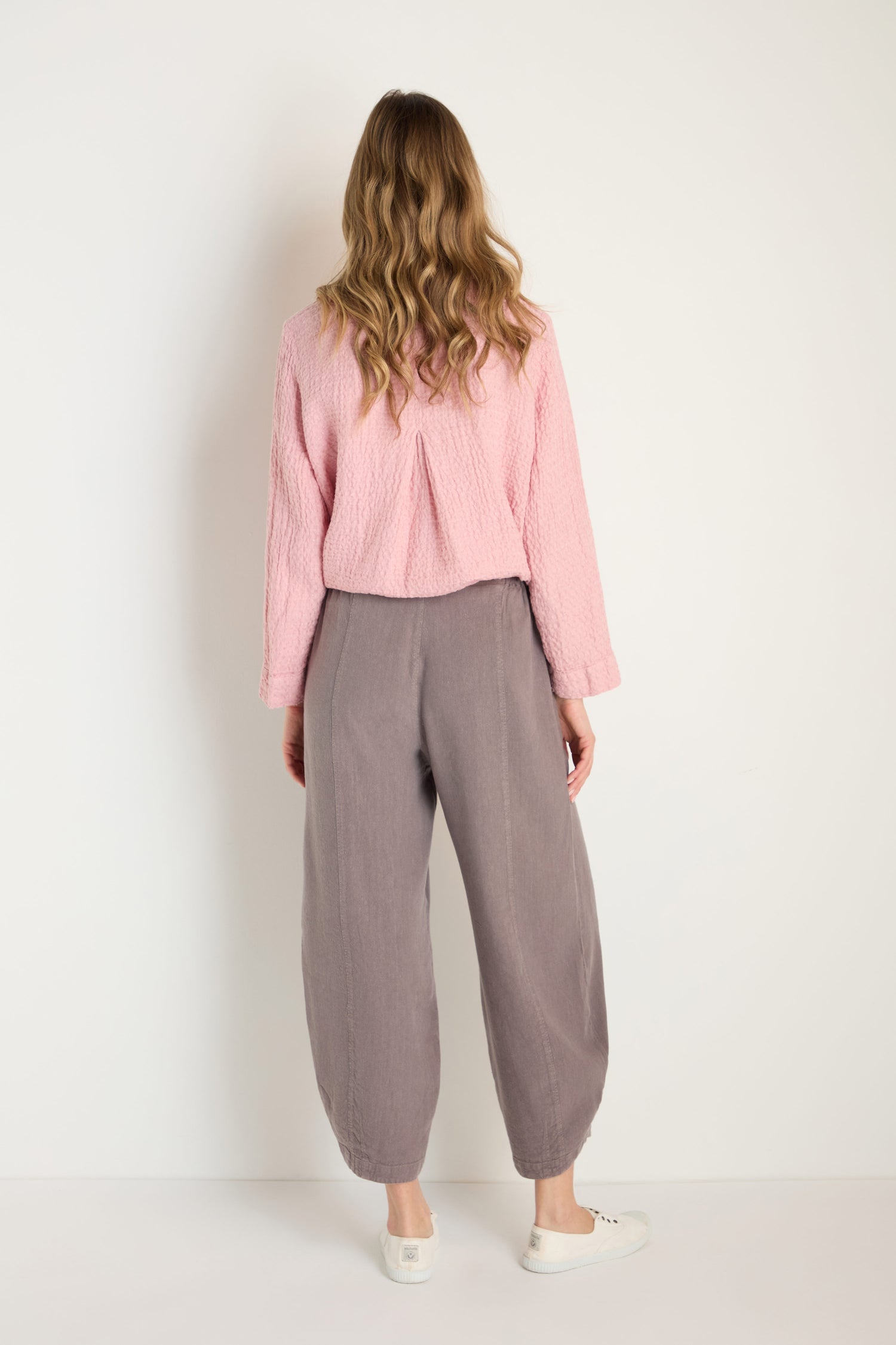 A person with long wavy hair, dressed in a pink textured top, Twisted Linen Bubble Trouser, and white shoes, stands with their back to the camera facing a white wall.