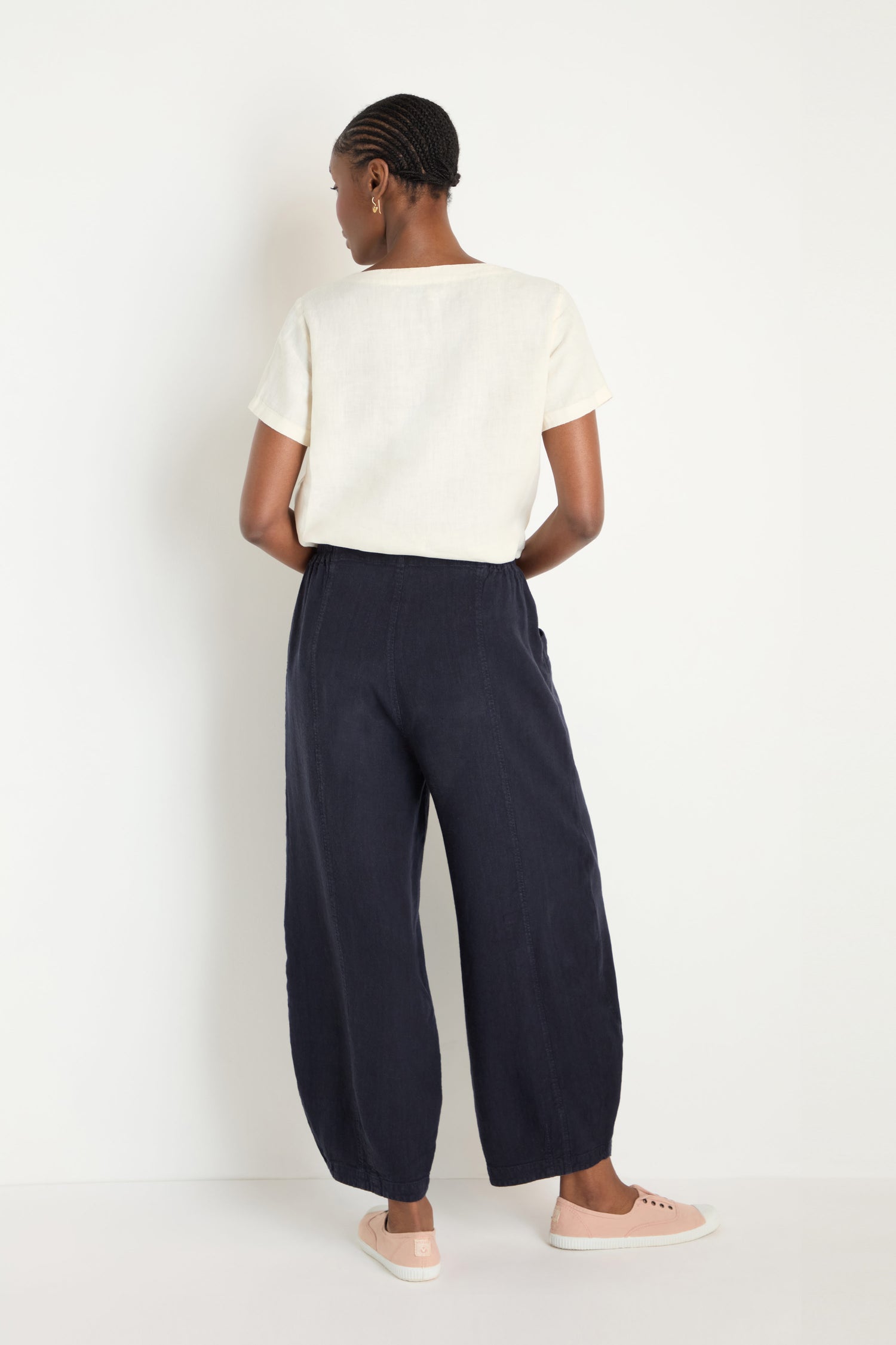 A person faces away from the camera, wearing a cream short-sleeve top, Twisted Linen Bubble Trouser in navy, and light pink slip-on shoes against a plain white background.