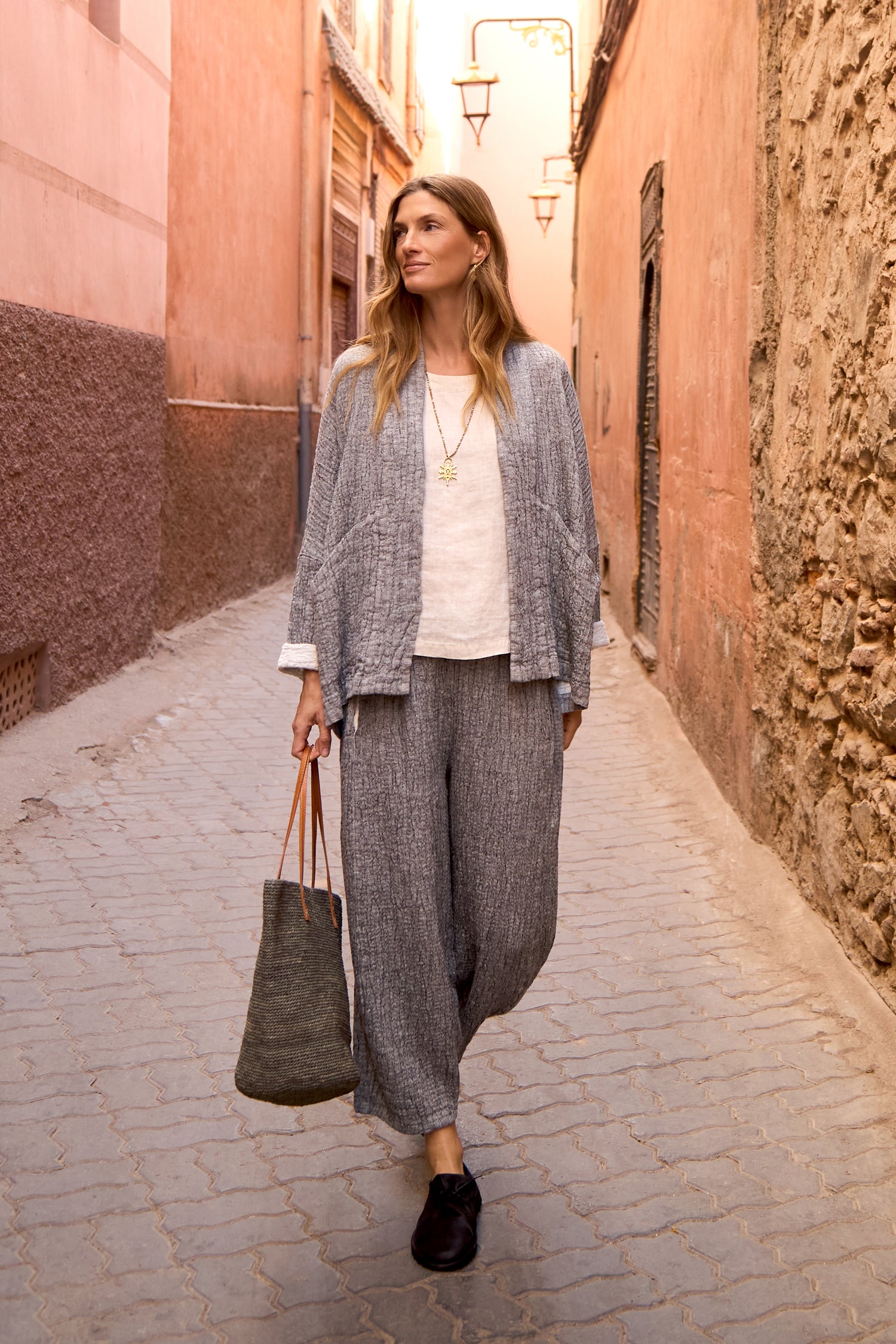 A woman wearing loose grey Crinkle Double Face Linen Bubble Trousers, a jacket, white top, and black shoes carries a tote bag as she walks down a narrow sunlit alley with pink walls.