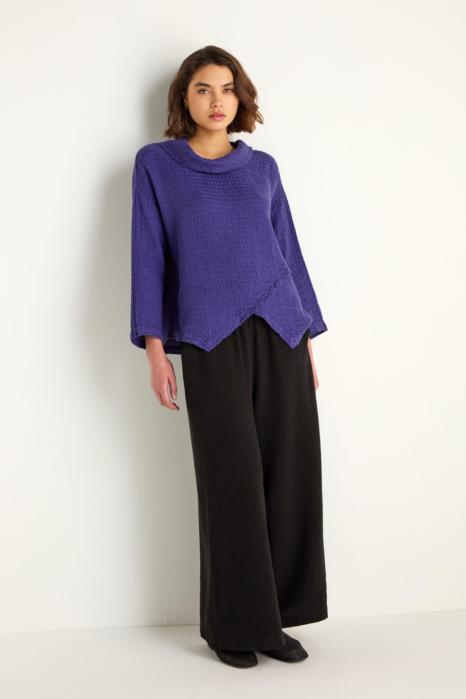 A person stands against a white wall wearing the Waffle Linen Boxy Cowl Neck Top in purple with a relaxed fit and asymmetrical hem, paired with wide-leg black pants.