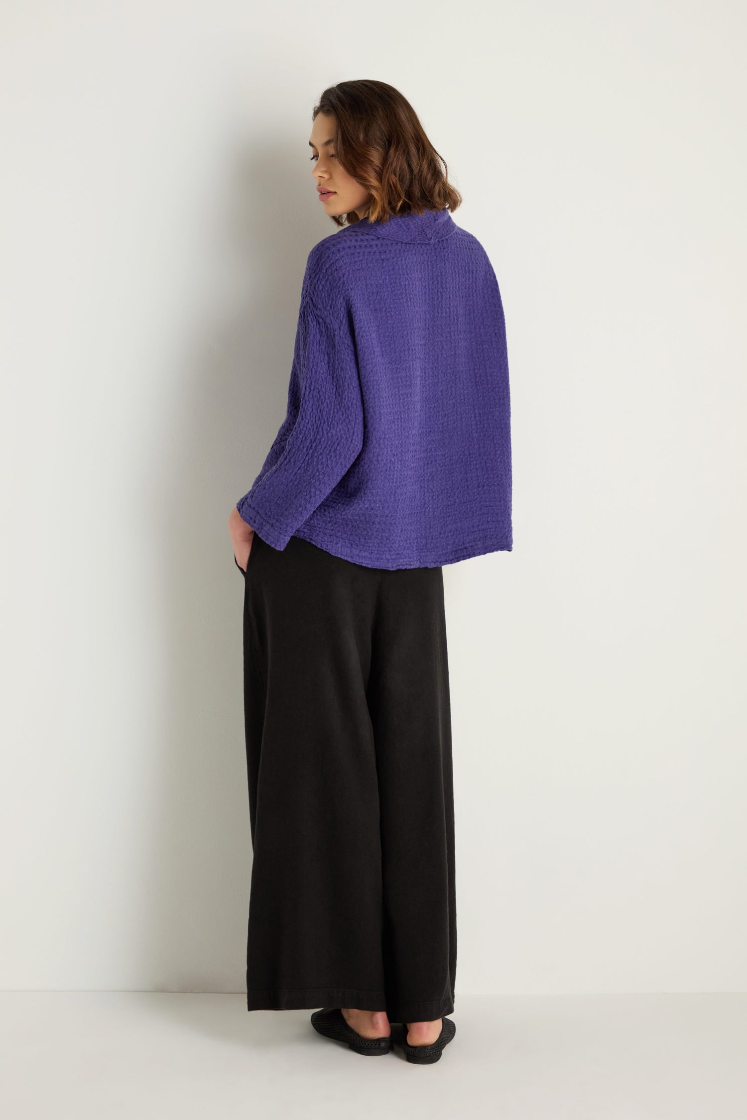 A person faces away from the camera wearing the Waffle Linen Boxy Cowl Neck Top in purple, paired with wide-leg black pants and black shoes, all set against a plain white background.