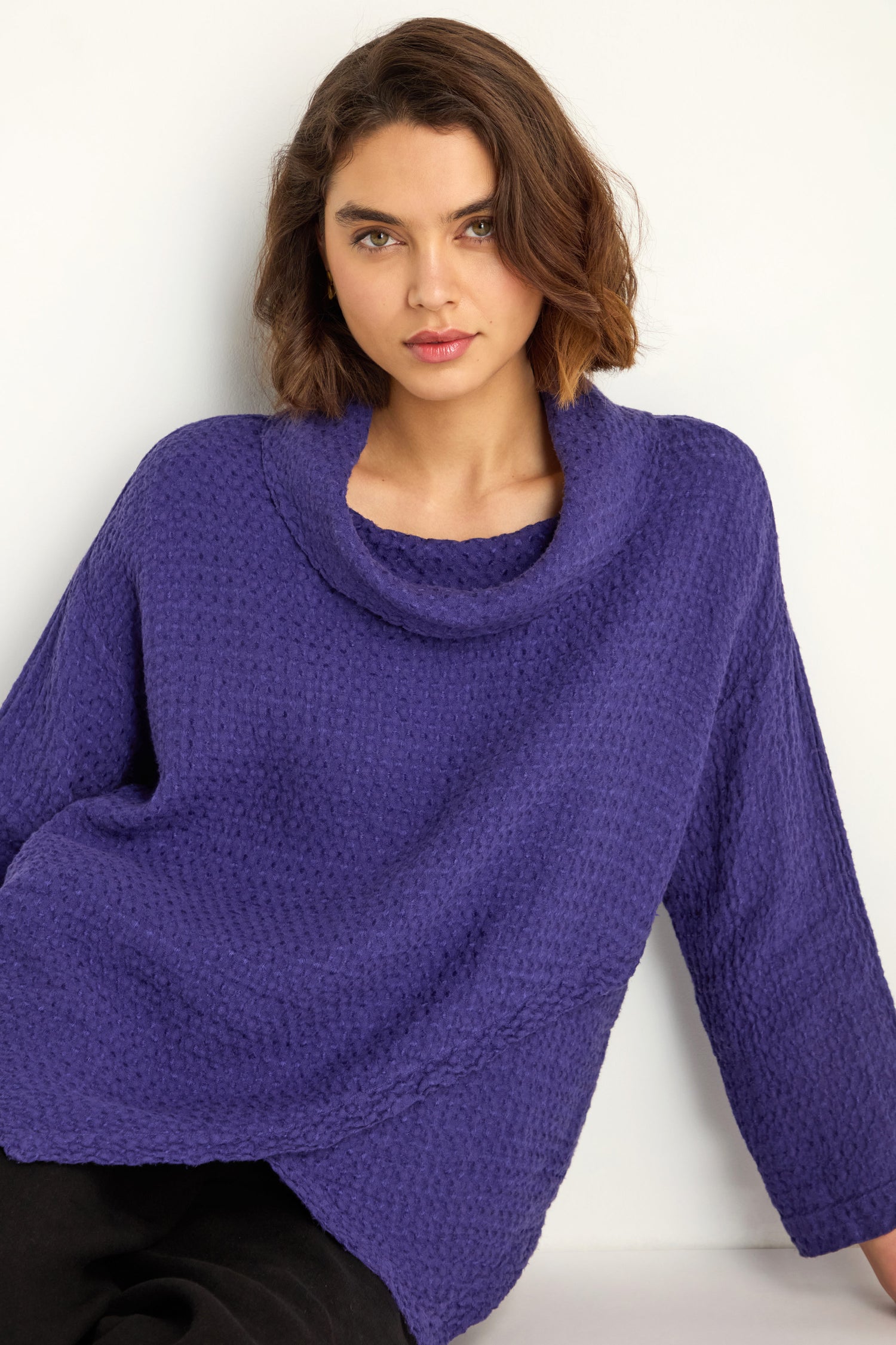 A woman with shoulder-length brown hair wears the Waffle Linen Boxy Cowl Neck Top in purple and black pants, seated against a plain white background.