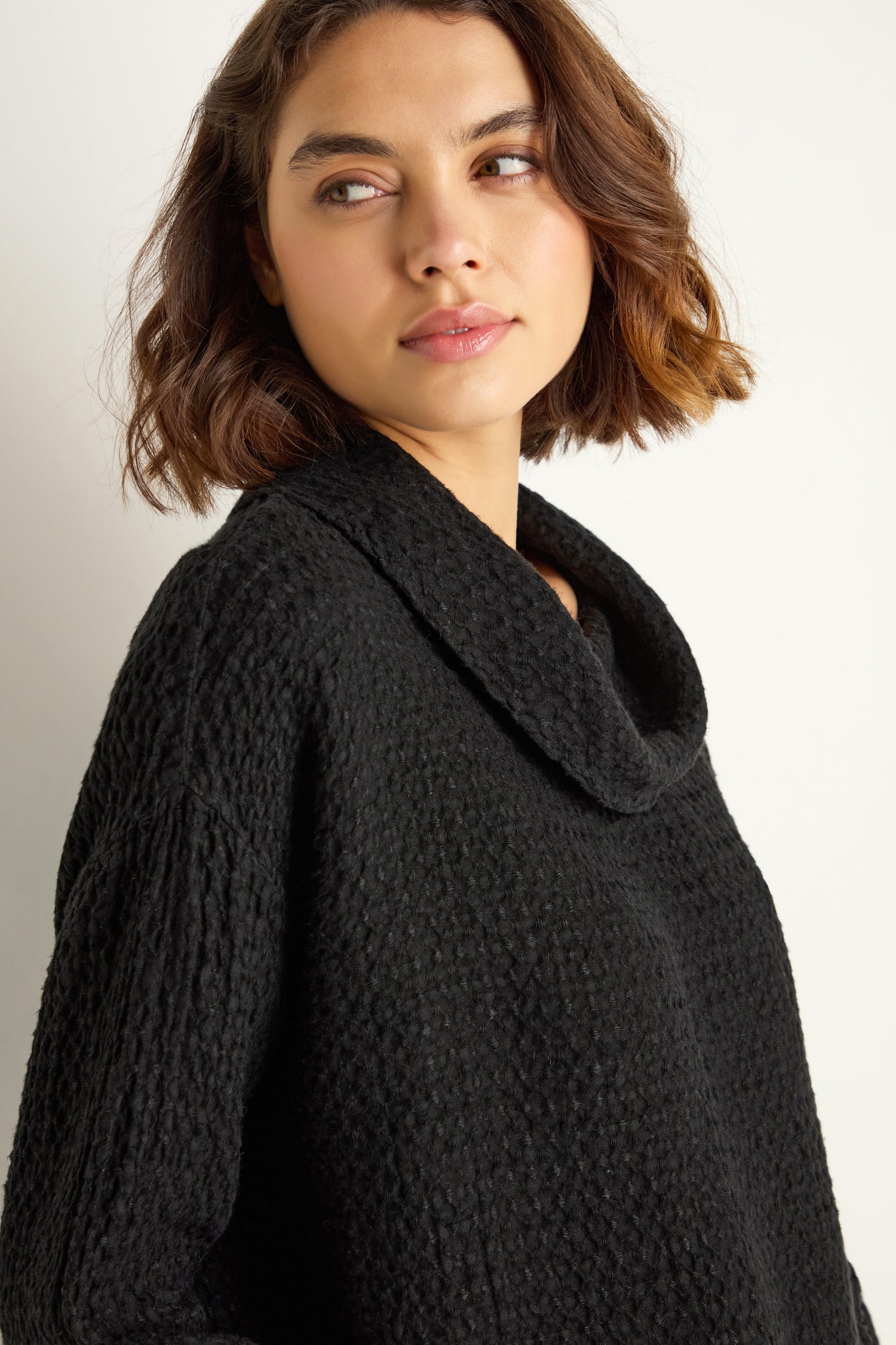 A woman with short brown hair wears the Waffle Linen Boxy Cowl Neck Top in black, relaxed fit, as she looks to her left against a plain light background.