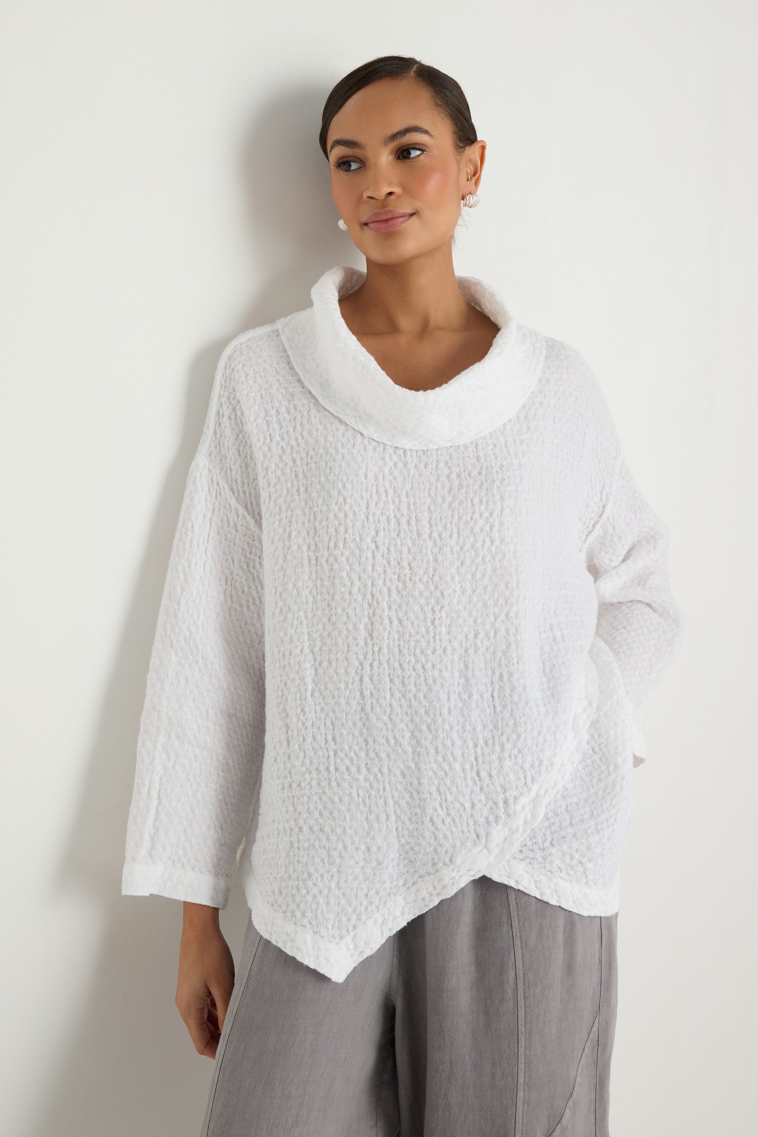 A person stands against a white wall wearing the Waffle Linen Boxy Cowl Neck Top and loose-fitting grey pants.