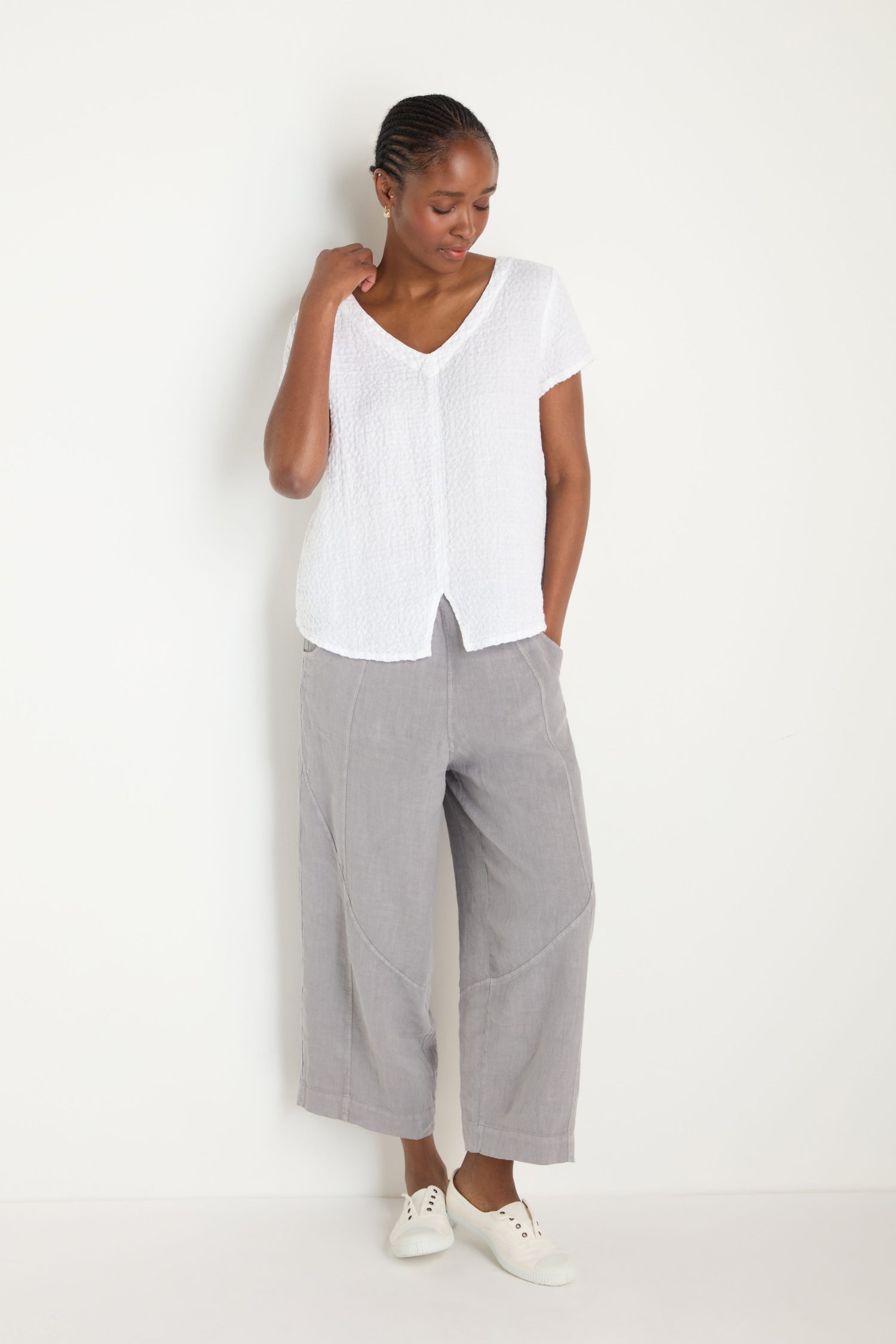 A woman stands against a white wall wearing a Waffle Linen V Neck Top, loose LAT6702 gray cropped pants, and white sneakers.
