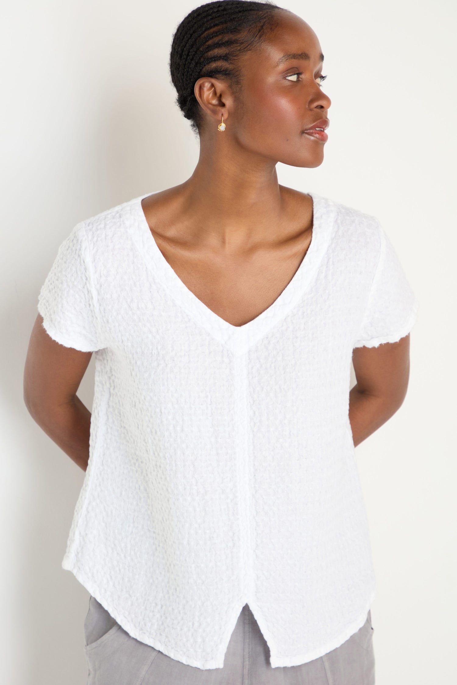 A woman in the Waffle Linen V Neck Top, a textured white short-sleeve design, and light grey pants stands looking to her right against a plain light background.