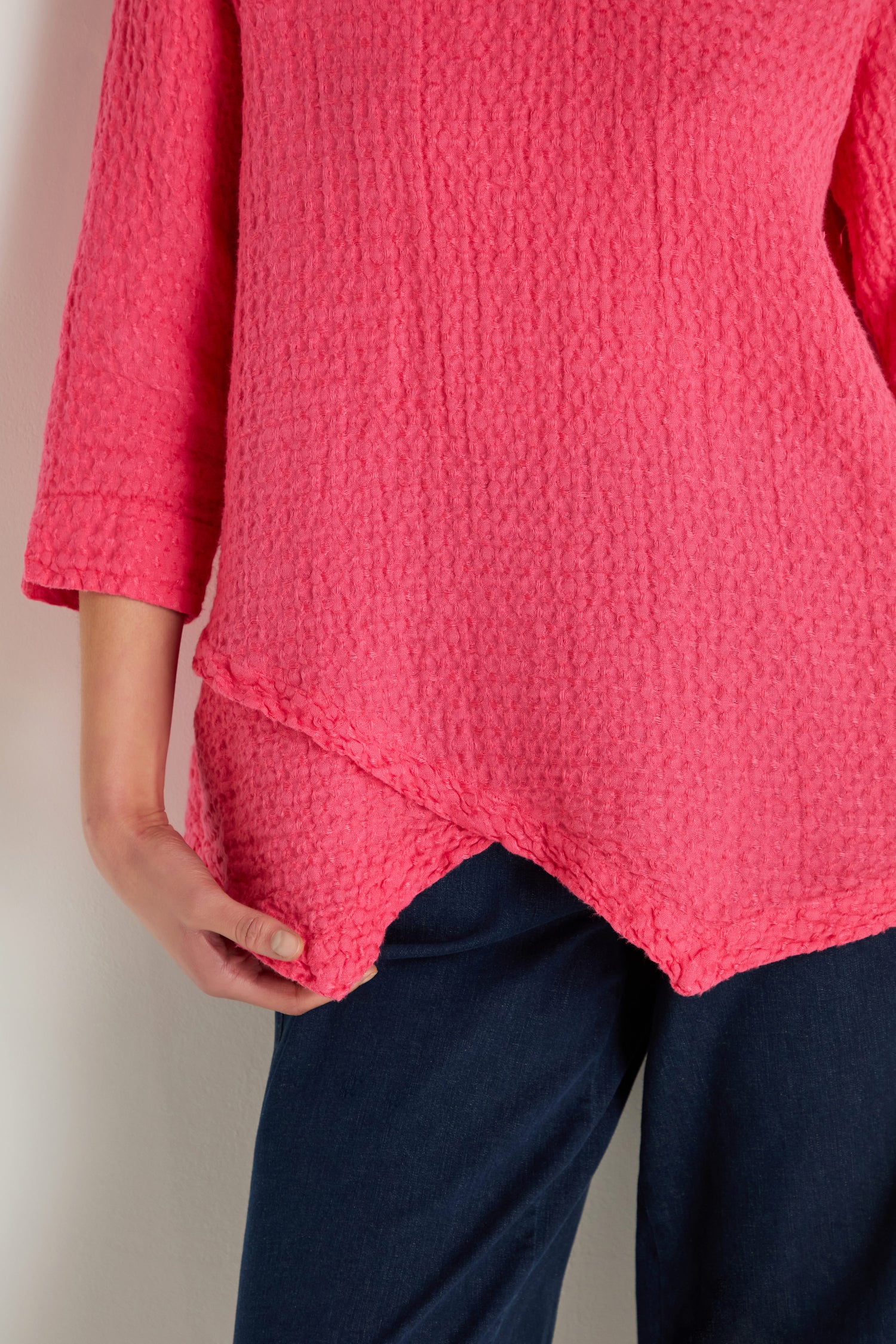 A person stands against a light-colored wall wearing the Waffle Linen Asymmetric Tunic in pink, paired with dark blue pants.