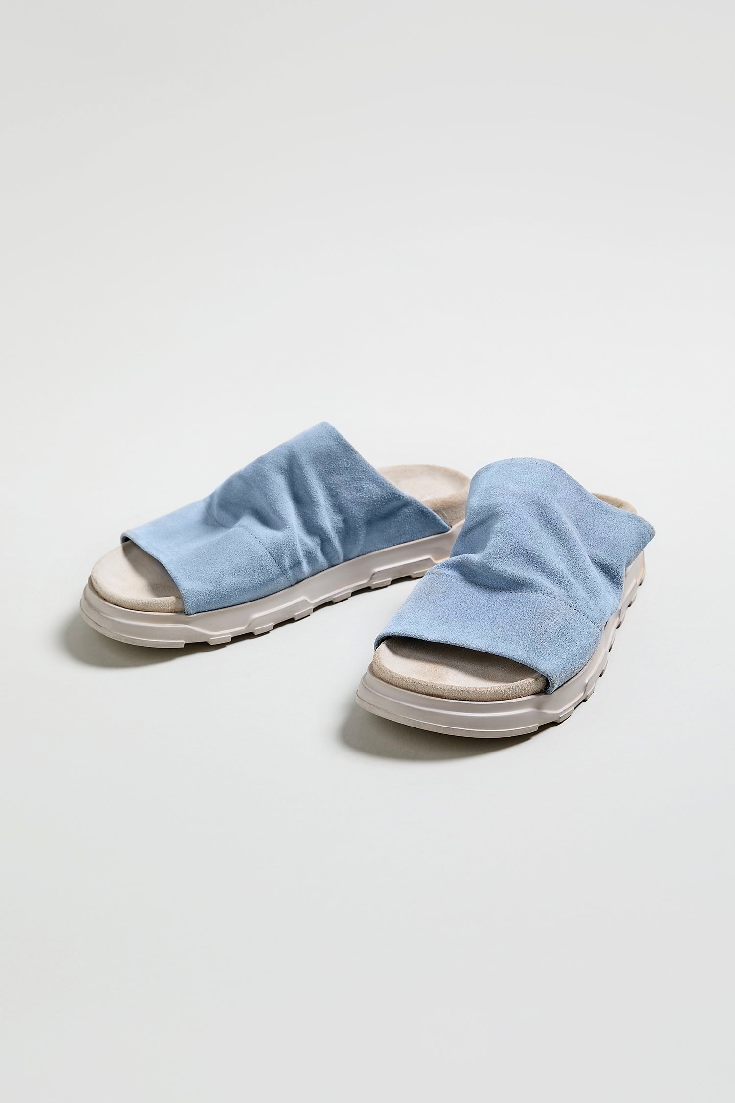 Step into summer with the Crushed Suede Sliders—light blue open-toe sandals featuring white soles, shown on a neutral background. A perfect SS26 wardrobe essential.