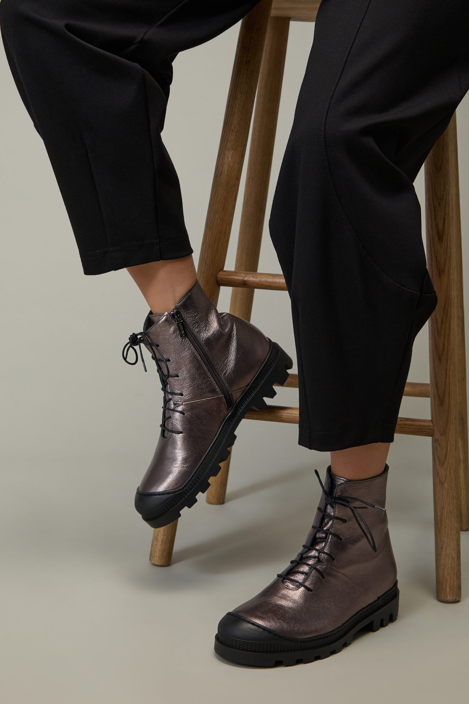 A person wearing Rose Metallic Lace Up Leather Boots with thick black soles, sitting on a wooden stool and dressed in black pants.