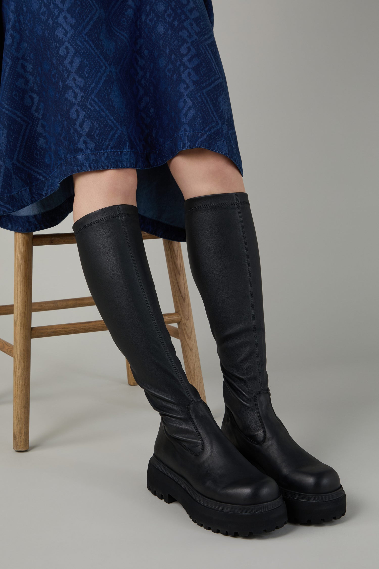 Person sitting on a wooden chair wearing a blue patterned skirt and striking Black Long Stretch Knee High Leather Boots with thick soles.