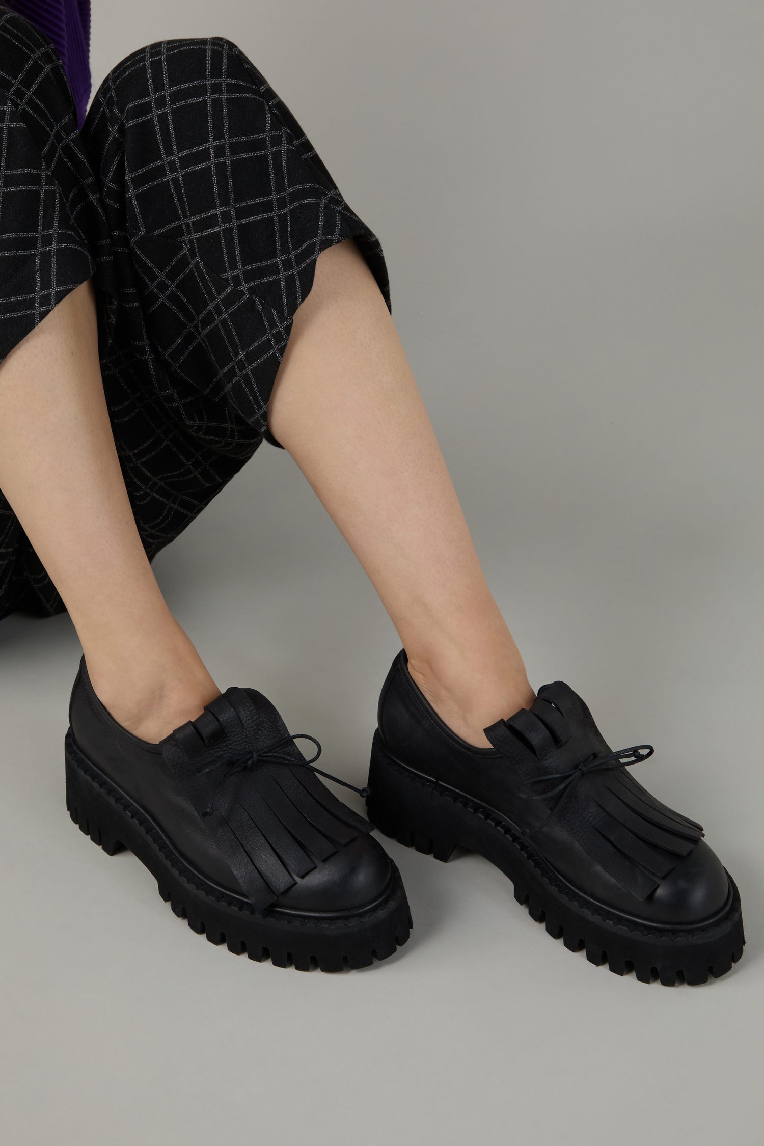Person wearing Fringe Top Leather Brogues with thick, rugged soles and checkered black pants, sitting on a plain grey background.