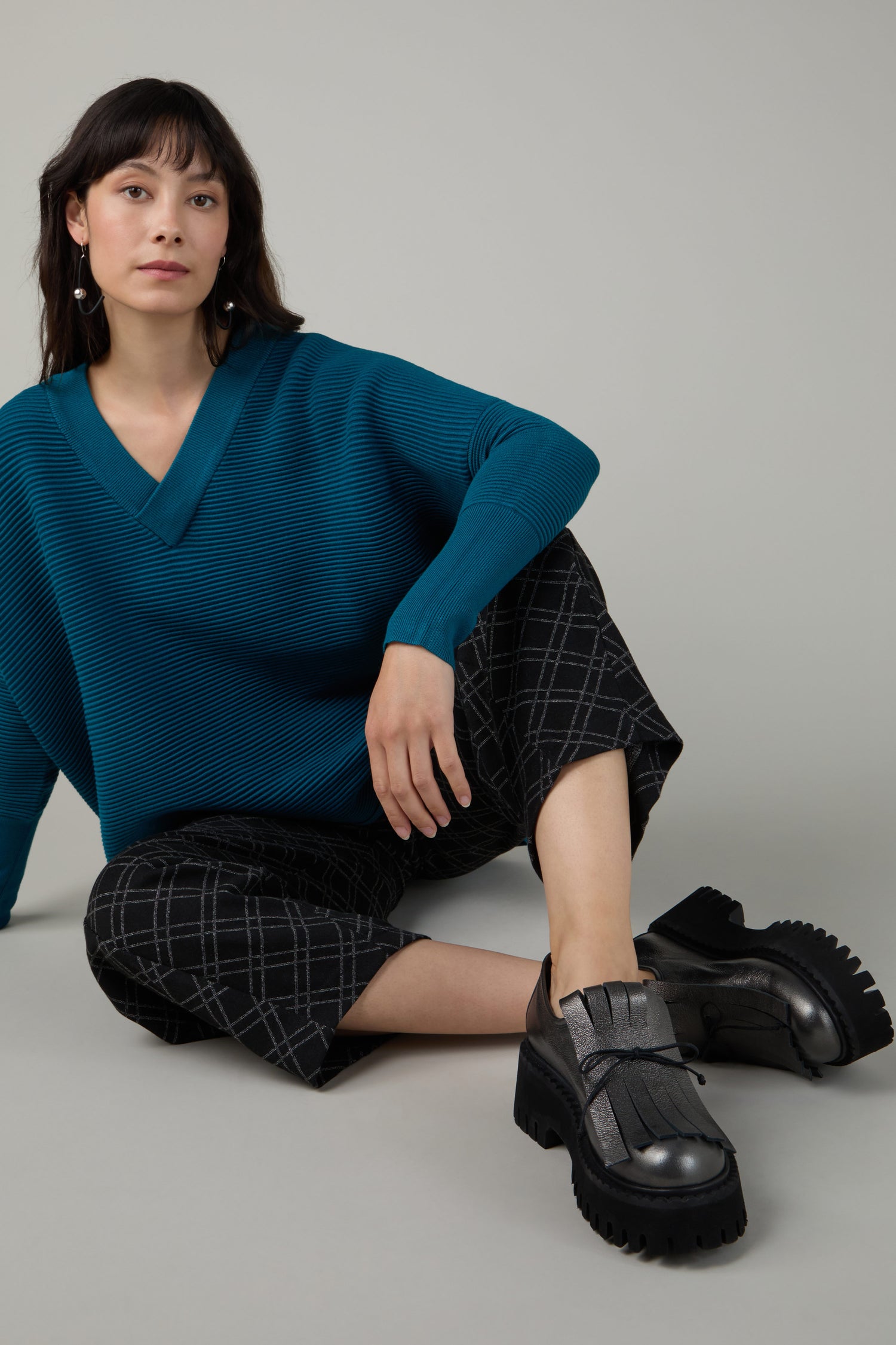 A woman with straight dark hair sits on the floor, wearing a teal V-neck sweater, plaid wide-leg pants, and Fringe Top Leather Brogues.