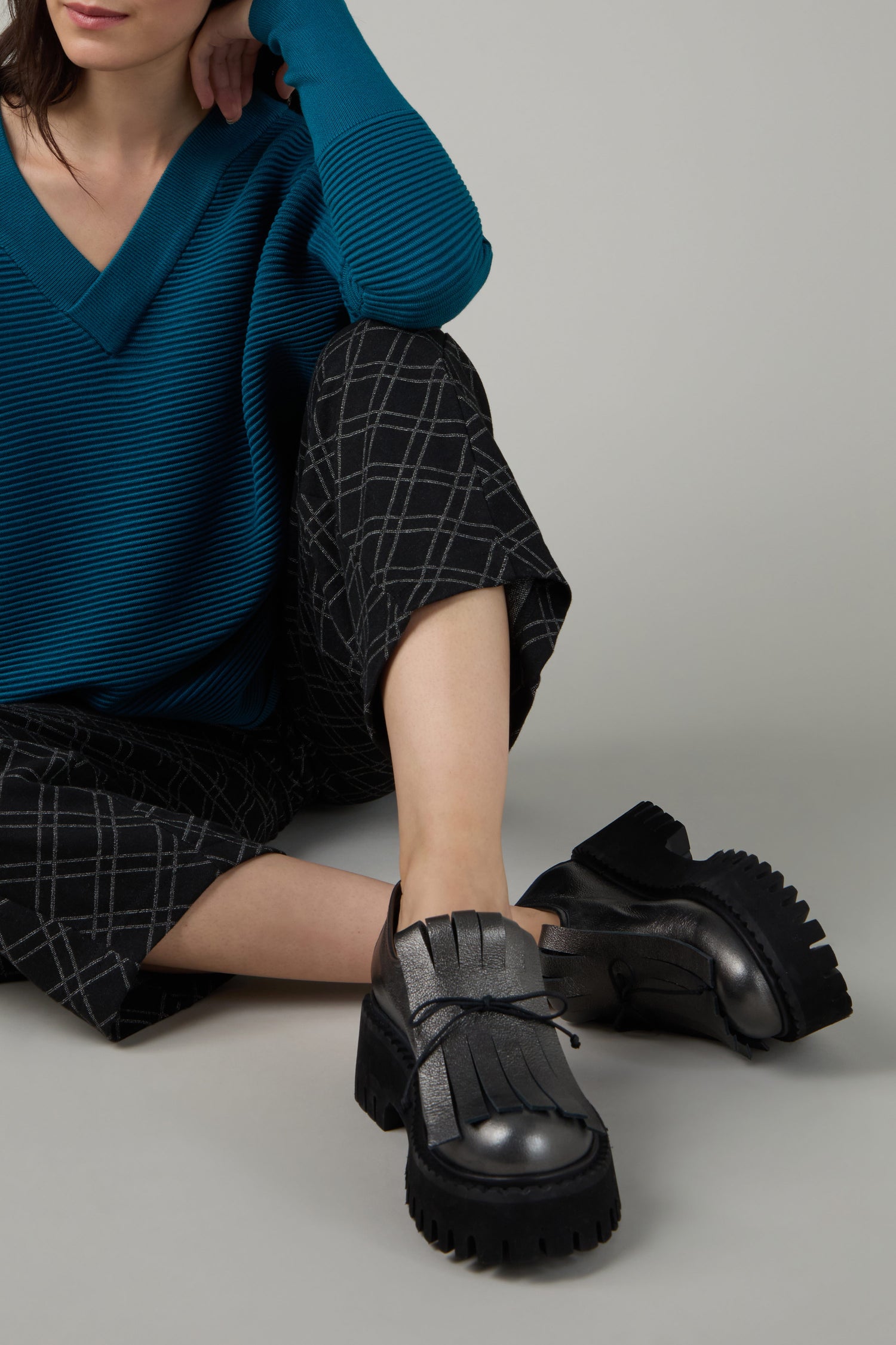 A person wearing a teal ribbed sweater, black and grey plaid pants, and Fringe Top Leather Brogues sits with one knee up and an arm resting on their knee against a plain background.