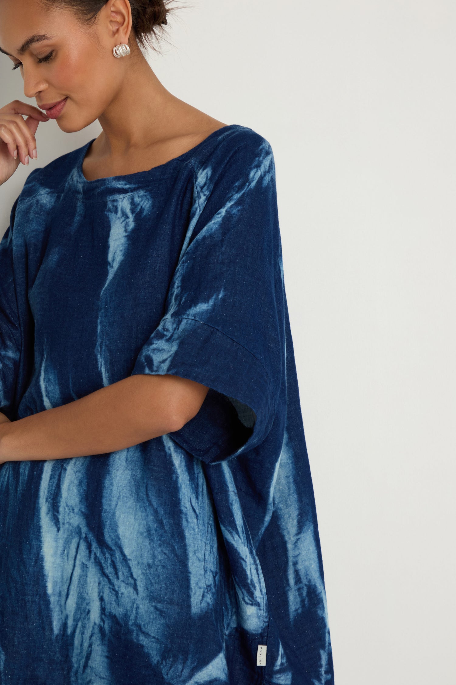 A woman models the Soft Denim Button-Back Tunic—loose-fitting and blue—standing against a white background, with one hand near her mouth and her other arm relaxed.