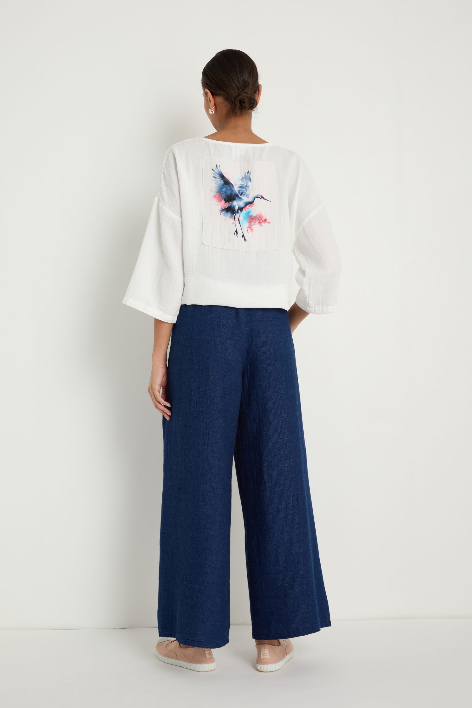 Woman standing against a white wall wears a loose white top featuring a crane and watercolor back design, Wide Leg Slit Trousers in dark blue, and beige flats.