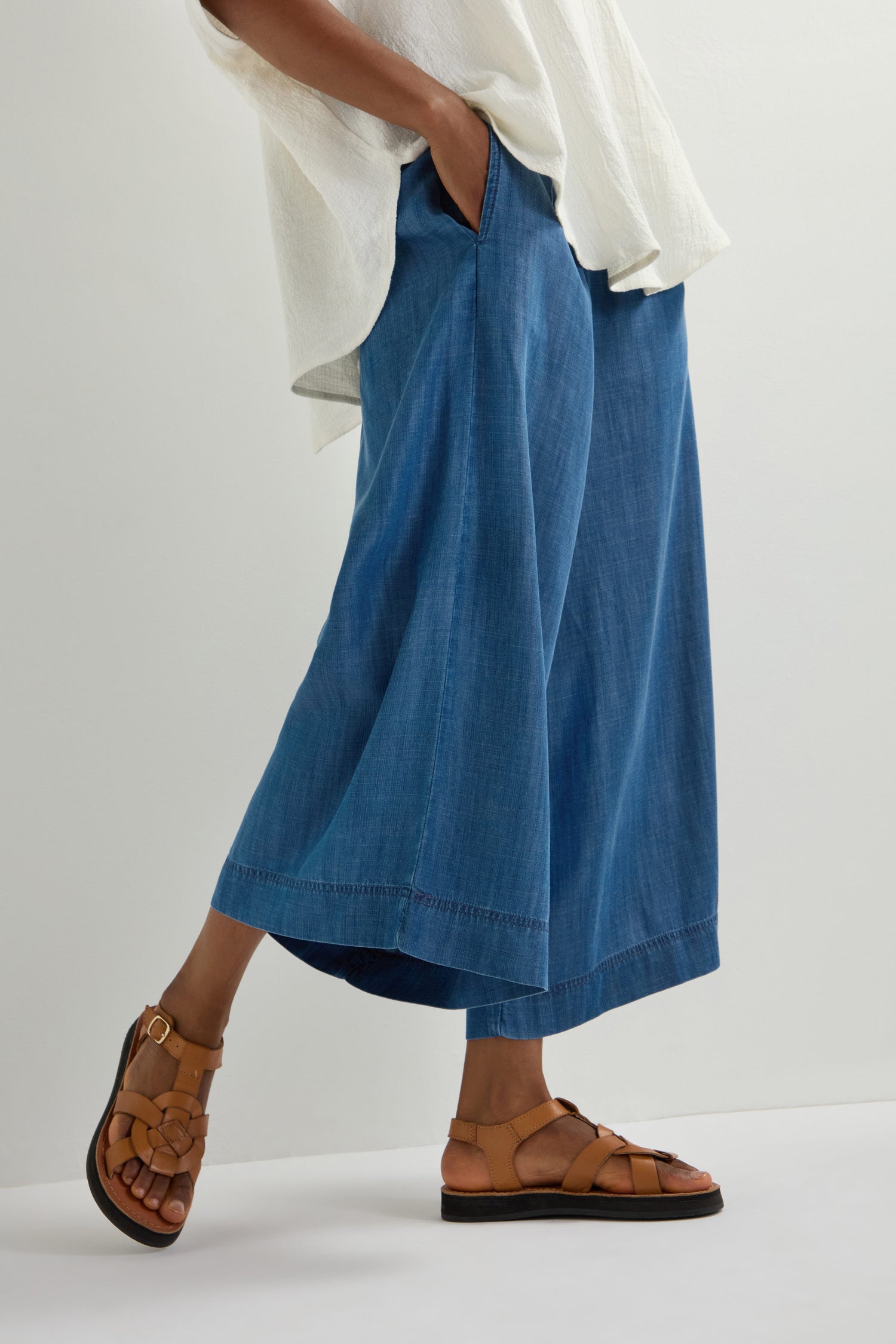 A person in a white shirt and wide-leg denim pants stands with one hand in a pocket, showcasing Tulum Leather Buckle Sandals against a plain background.