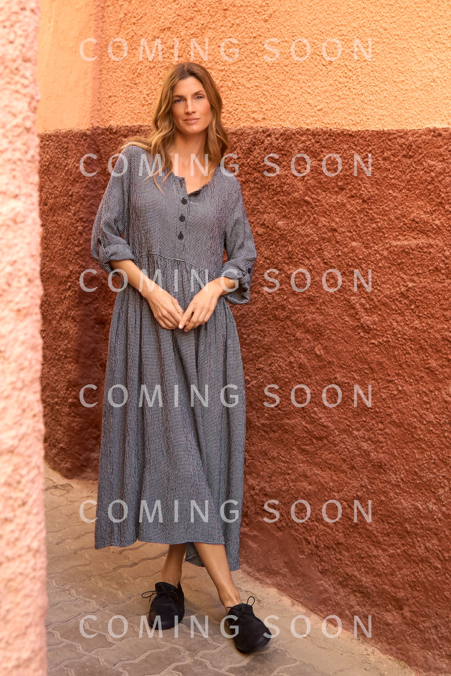 A woman stands outdoors by a two-toned wall wearing the Micro Gingham Bubble Dress with black slip-on shoes. Text overlay reads "COMING SOON.