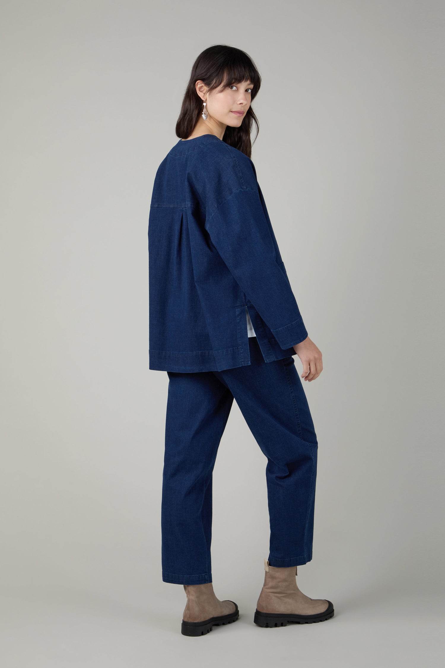 A woman stands facing slightly away, wearing the Stretch Denim Boxy Jacket in a dark indigo wash with matching pants and light brown ankle boots against a plain background.