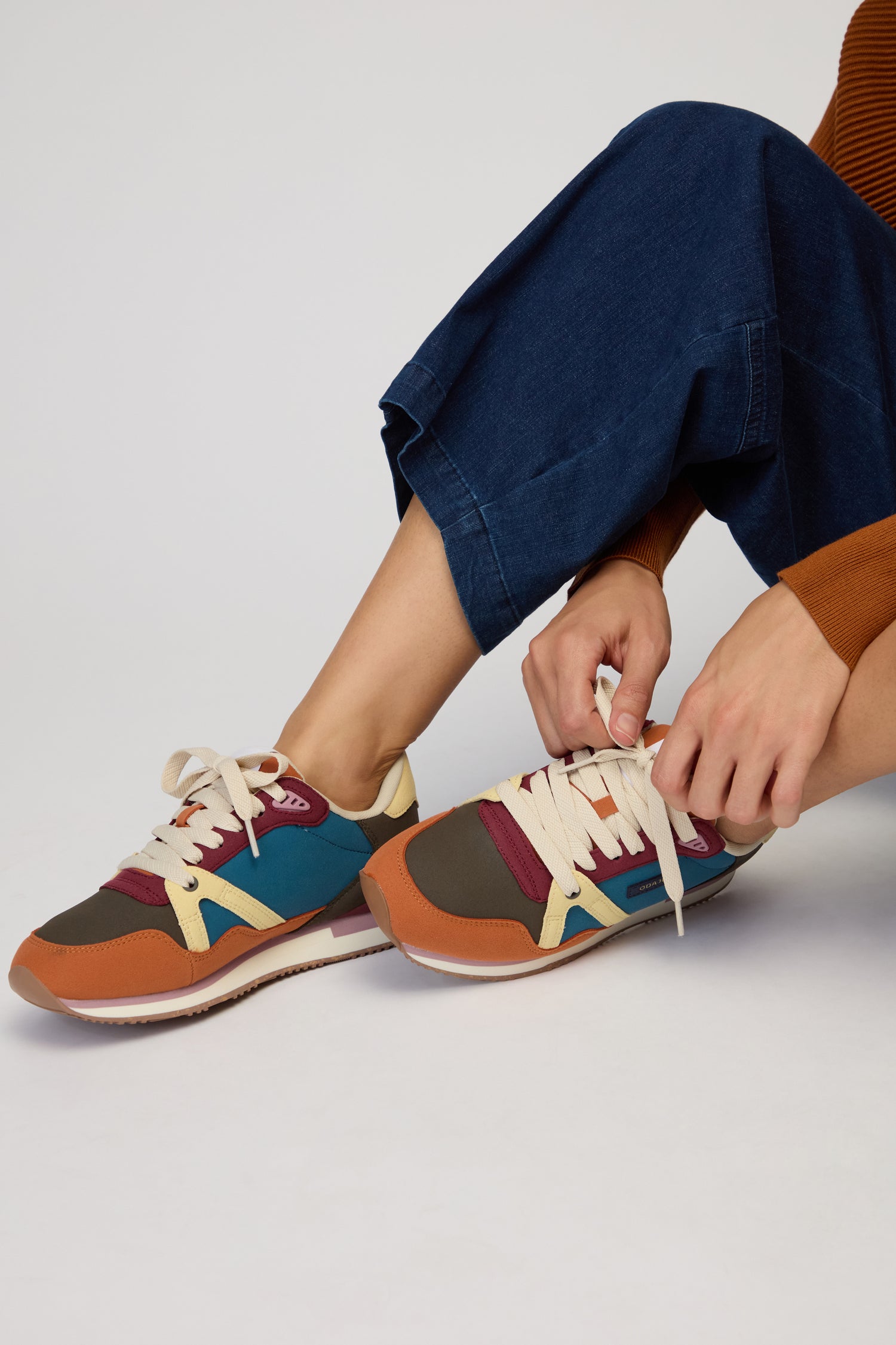 Wearing blue jeans and a brown sweater, the person sits tying the laces of Colour Pop Andree Trainers—vegan shoes featuring bold colour blocking in red, teal, beige, and brown.