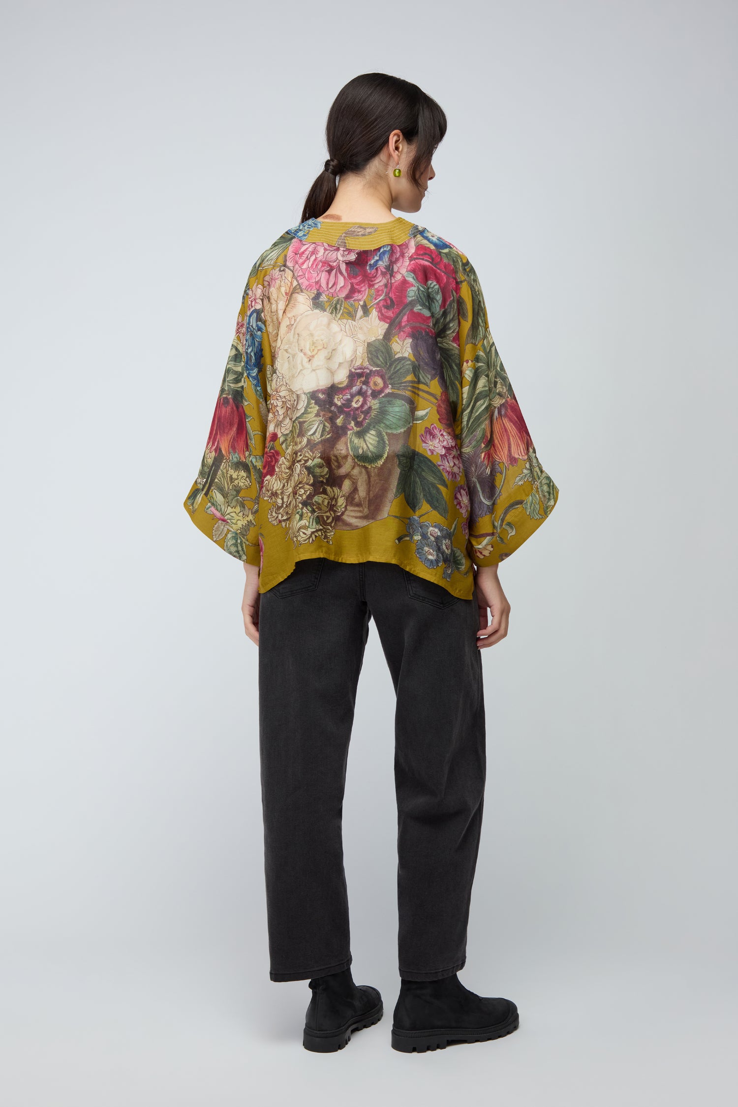 A woman stands with her back to the camera in a bold yellow floral Primula Kimono, paired with black pants and black shoes against a plain light gray background.