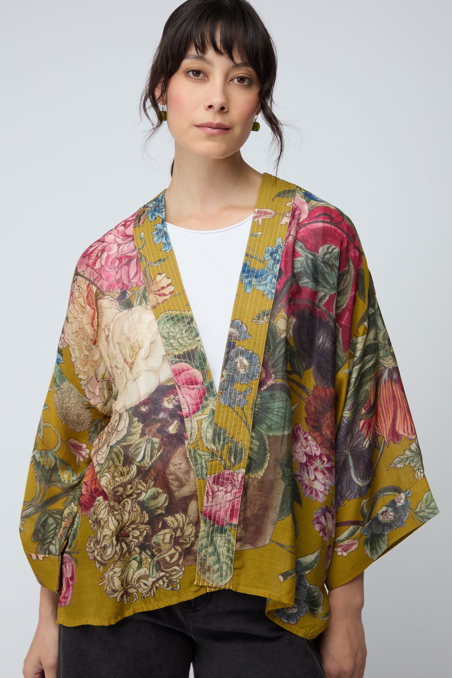 A woman stands against a plain light background wearing a white top and the Primula Kimono, which features an artistic floral print.