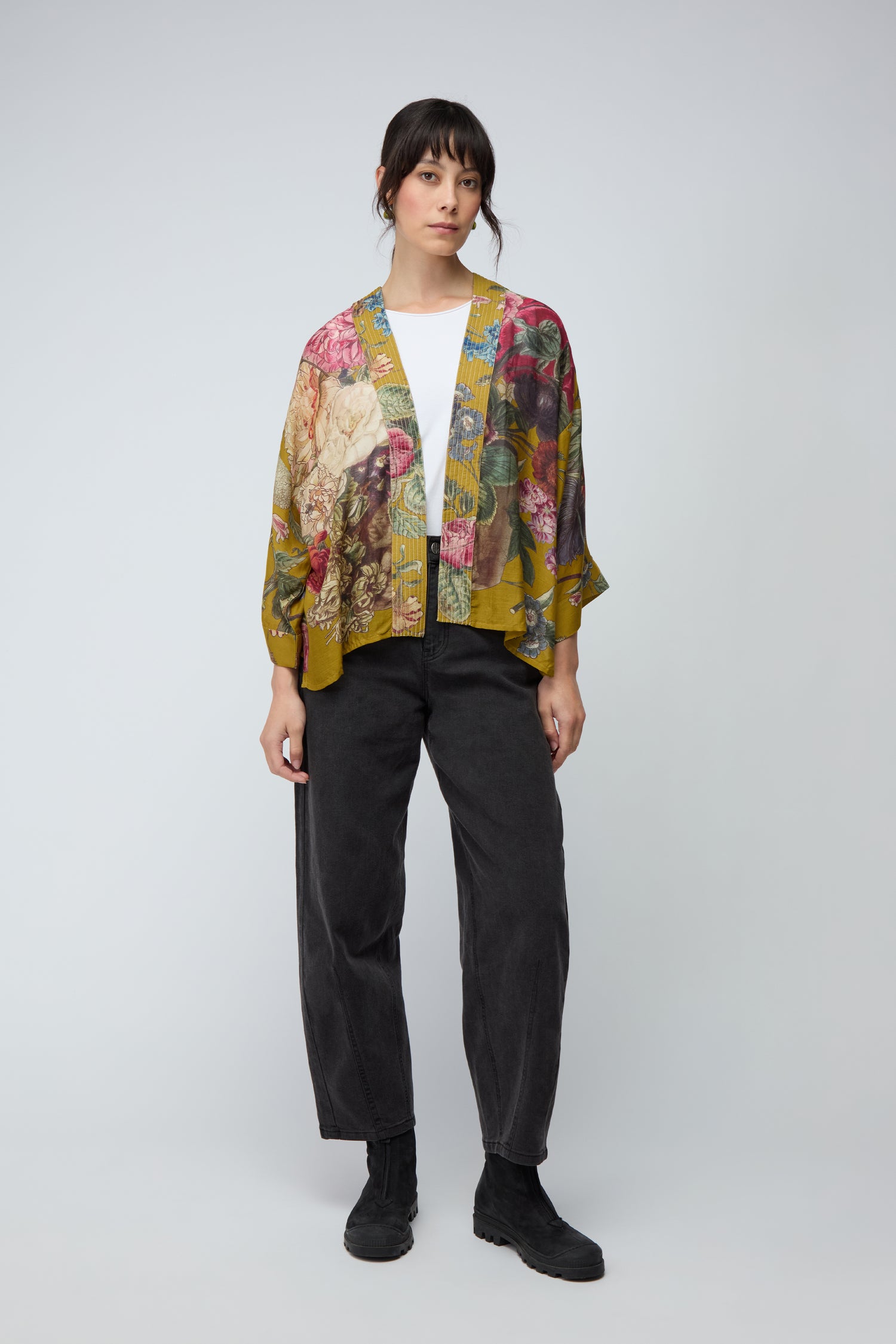 A person stands against a plain background, wearing a white shirt, loose black pants, black boots, and the Primula Kimono—a floral print cardigan that adds a vibrant touch to the look.