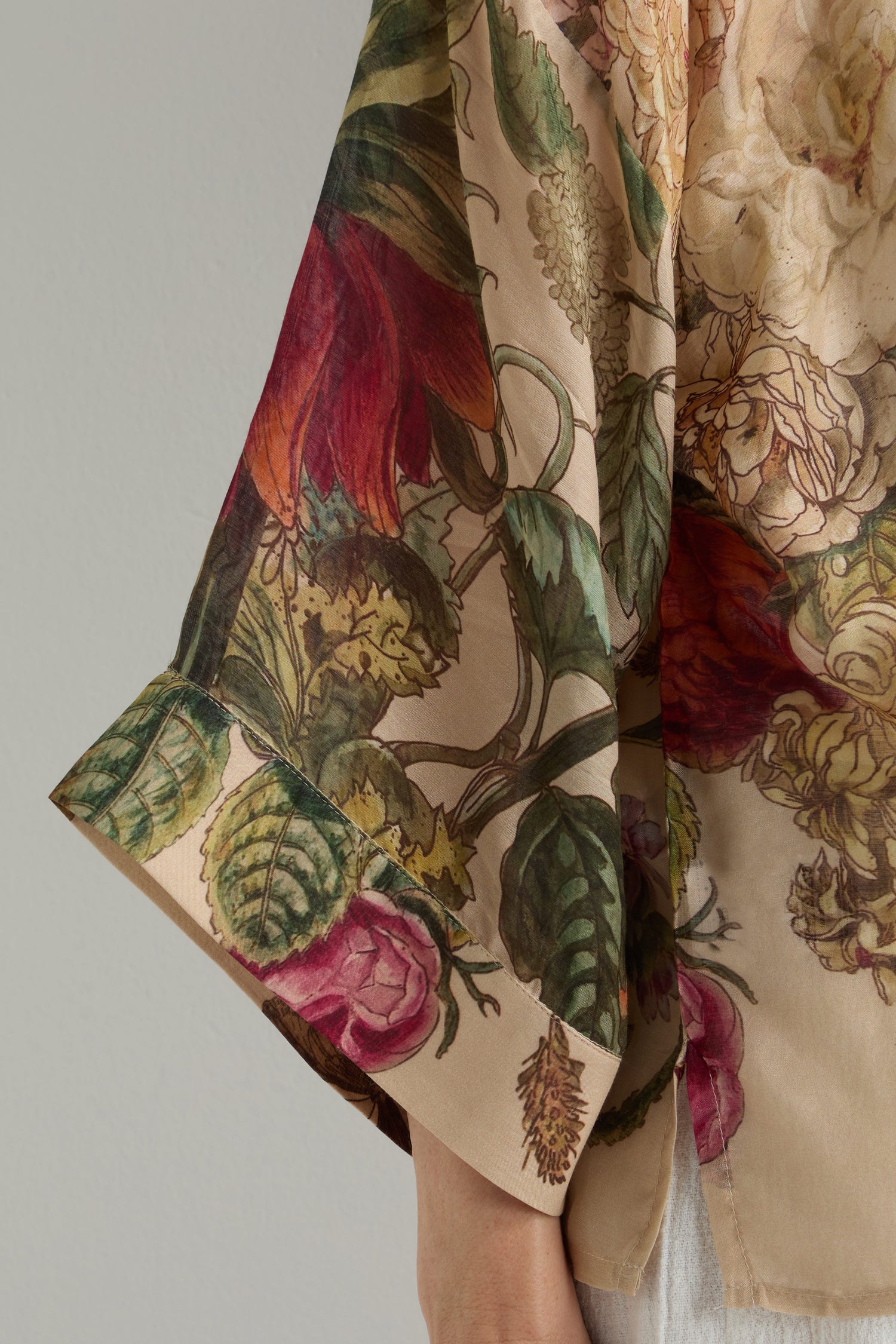 Wearing the Primula Kimono, a beige, sheer long-sleeve top with red, pink, and green floral designs, exudes artistic flair inspired by Dutch Old Masters.