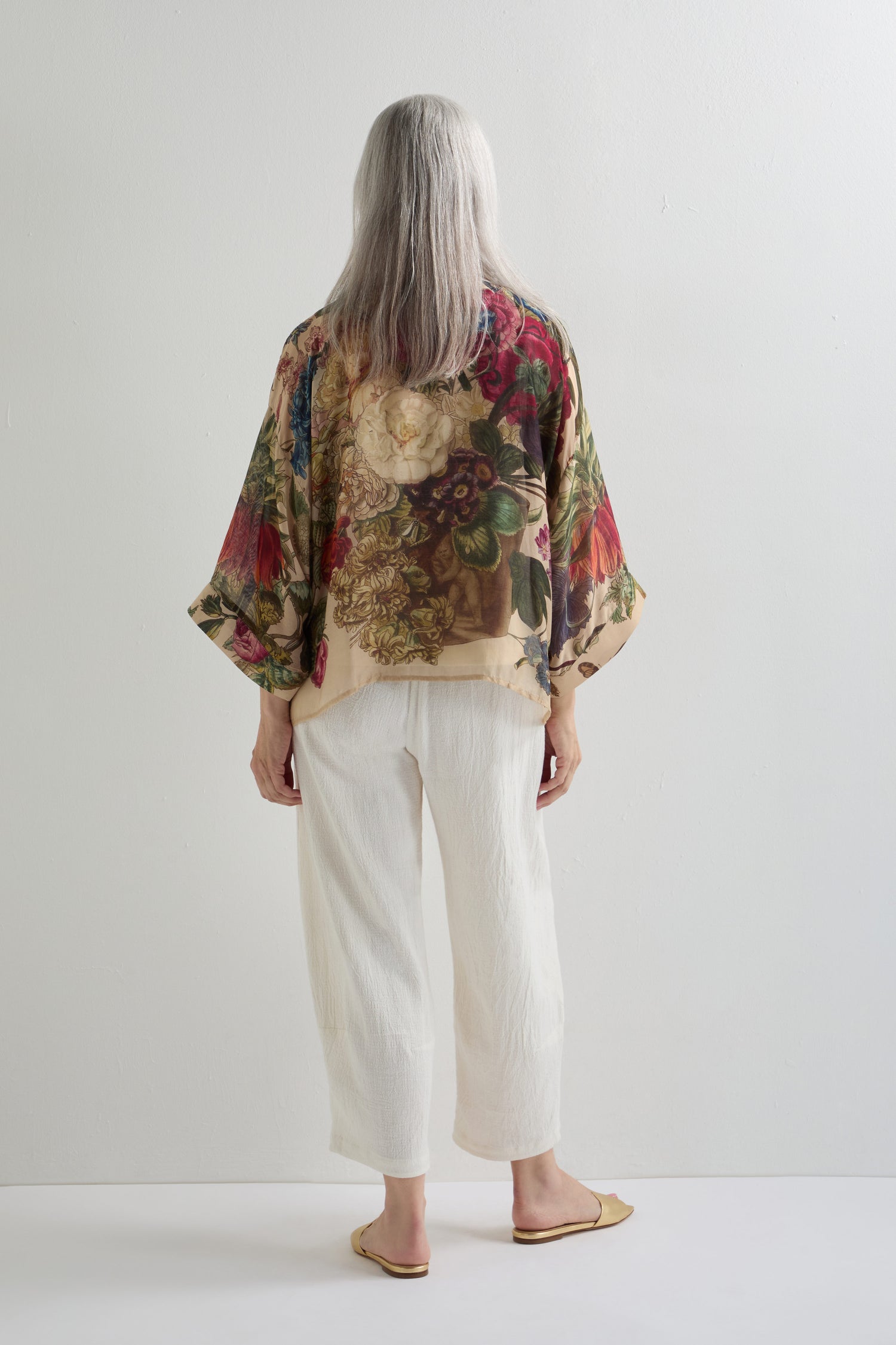 Wearing the Primula Kimono, a person with long gray hair faces a white wall, styled with white pants and beige flats for an artistic look.