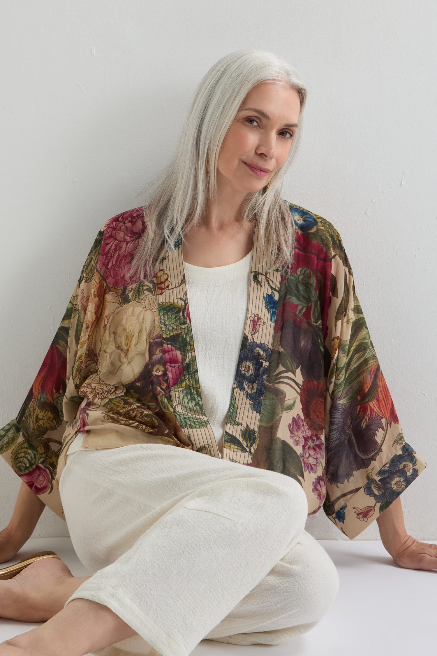 A woman with long gray hair sits on the floor against a white wall, wearing a white outfit and the Primula Kimono, which adds an artistic flair to her serene pose.