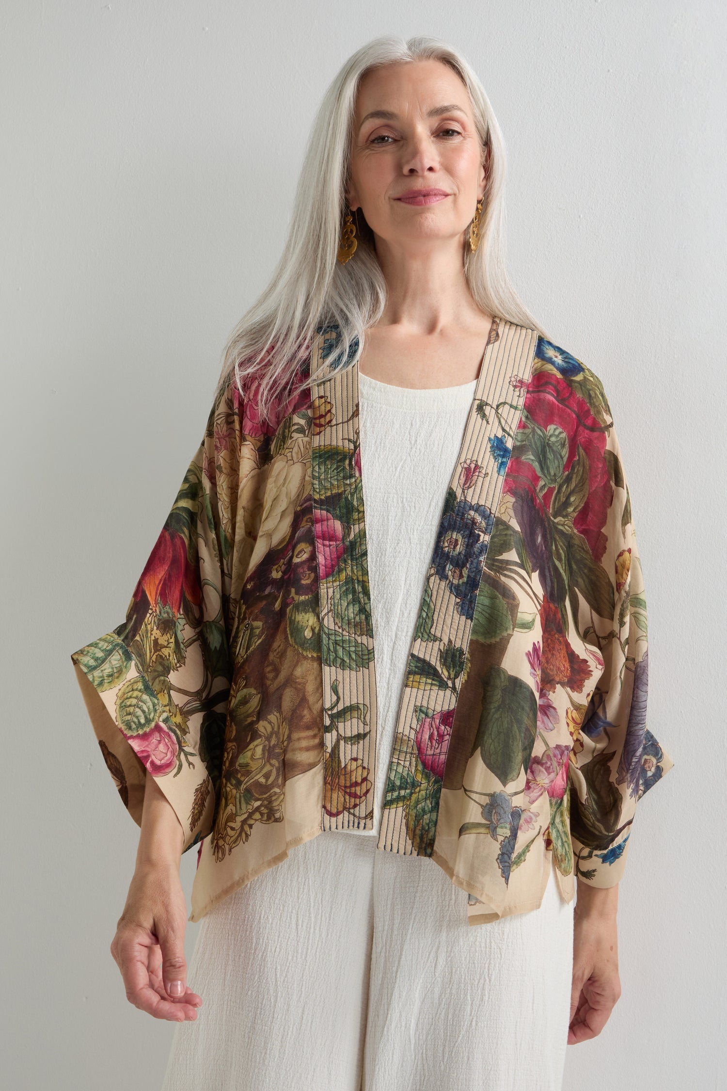 An older woman with long gray hair wears the Primula Kimono over a white top and pants, standing against a plain light background, exuding artistic flair reminiscent of Dutch Old Masters portraits.